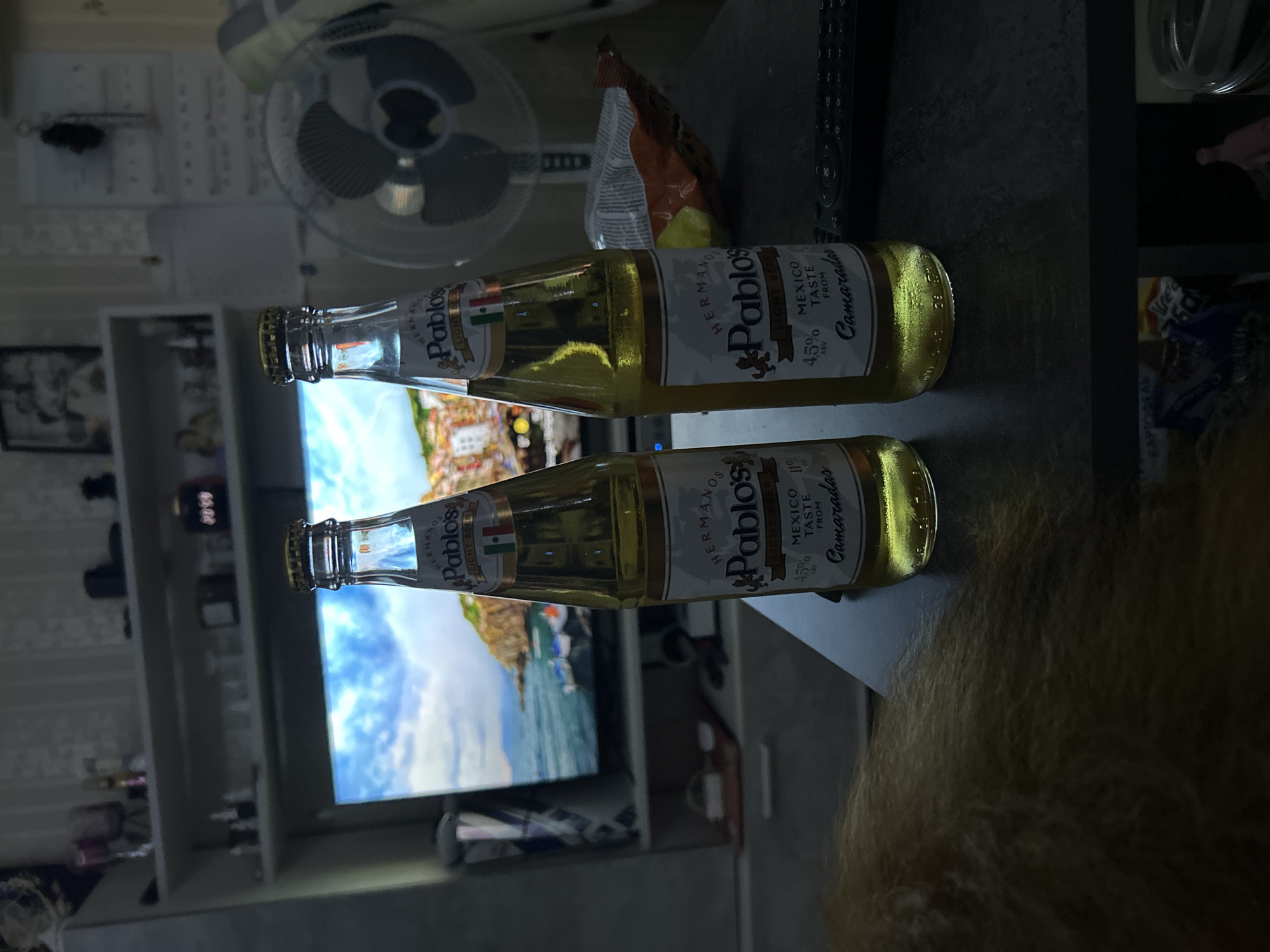 The image shows two bottles of "Hermano Pablo's" light beer, likely resting on a dark surface. A fan is visible in the background, along with a television screen displaying a coastal scene. Other objects such as a bag of chips, remote control, and shelf with various items are also present in the background. The image is shot with a slight rotation. No specific information indicates the location is in Khimki, Russia.