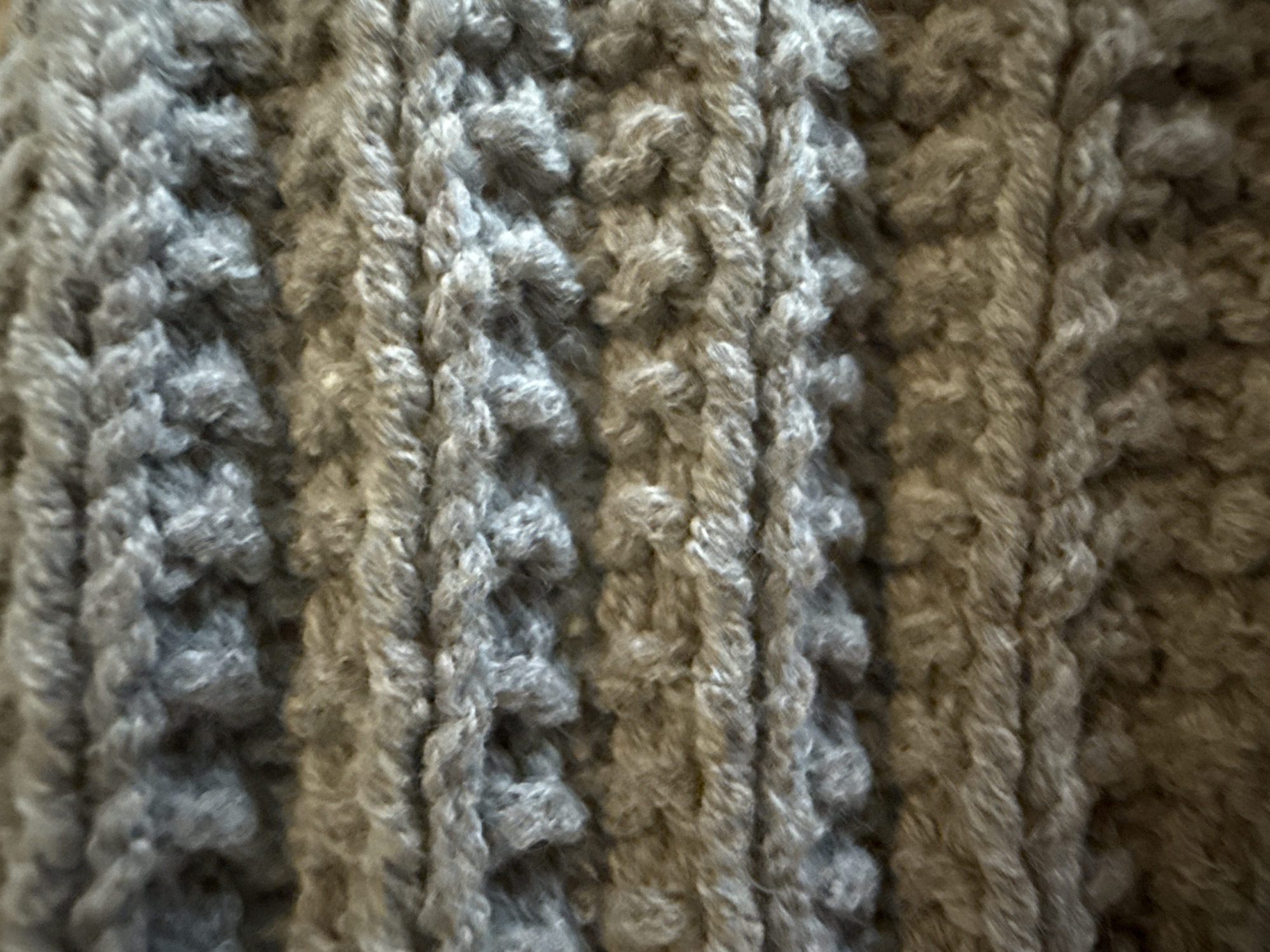 This is a close-up, static shot of a grey, textured fabric, likely a knitted material. The texture is characterized by raised, ribbed patterns that run vertically, creating a sense of depth and dimension. The fibers of the fabric appear soft and slightly fuzzy, suggesting it could be wool or a similar synthetic blend. The lighting casts subtle shadows within the grooves of the knit, further accentuating the texture. There are no people, objects, or scenes visible, and no activity or event is taking place. The image is a purely abstract representation of the fabric's surface. There is no visible text. The location context of Dolgoprudny, Russia, does not provide any visual cues within the image itself.