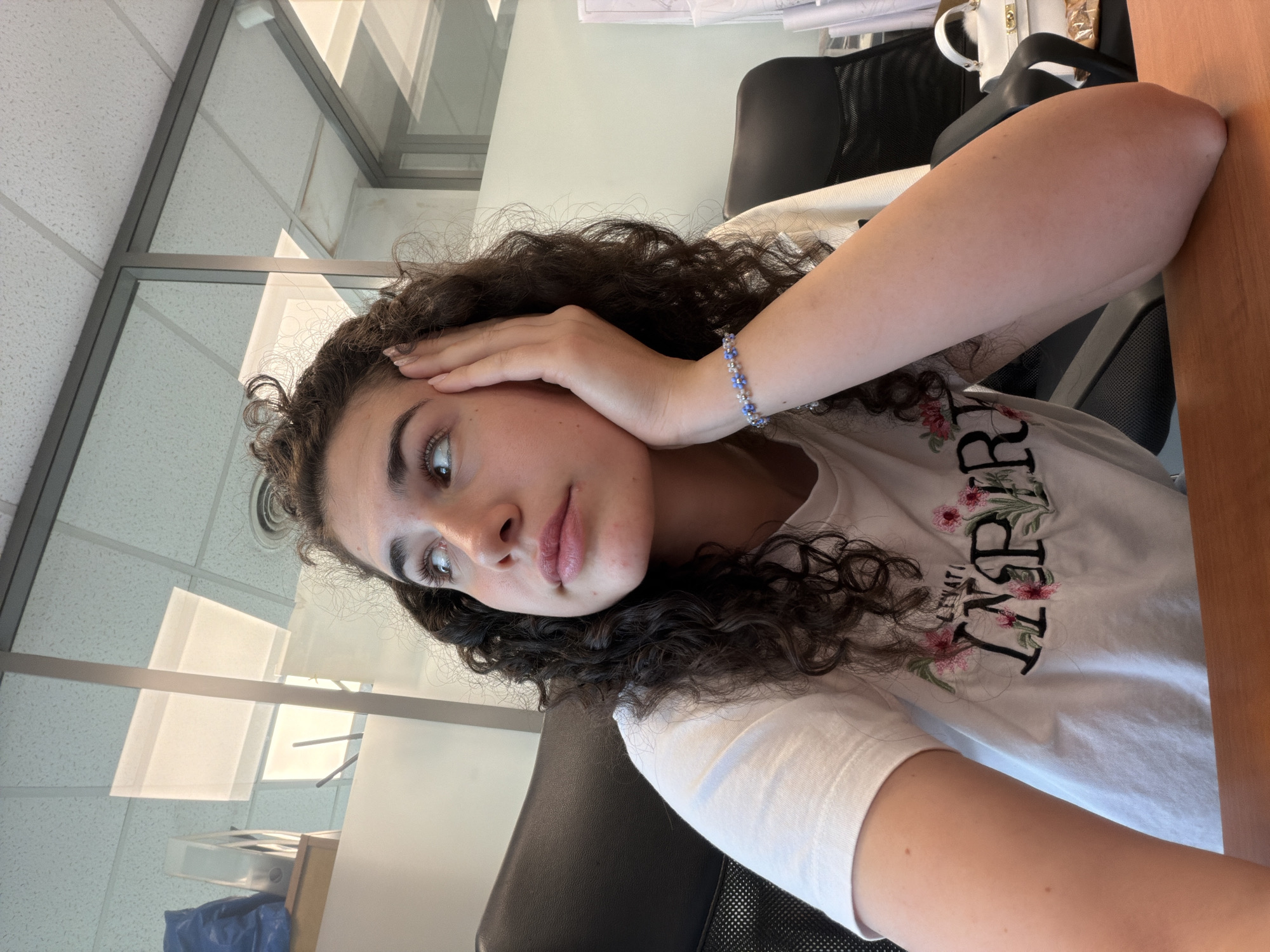 A young woman with curly brown hair is sitting at a desk in an office environment. She is looking upwards and to her left with her right hand resting on her cheek. She is wearing a white t-shirt with a floral design and the word "INSPIRE" embroidered on it, and a blue beaded bracelet on her right wrist. The office has glass partitions and fluorescent lighting. A black office chair is visible behind her, and a white handbag is on a surface to the right. The scene appears to be in Sofia, Bulgaria.