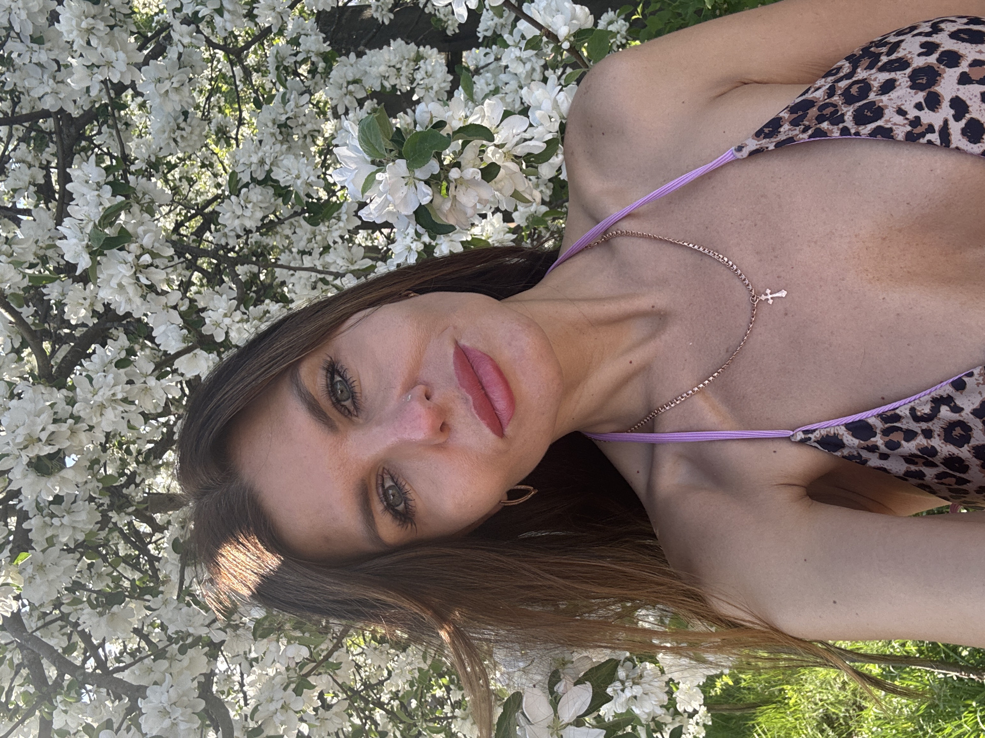 A woman is posing for a picture. She is wearing a leopard print bikini top and a necklace with a cross. She is standing in front of a tree with white blossoms and green grass. The location is Dubovaya, Russia.