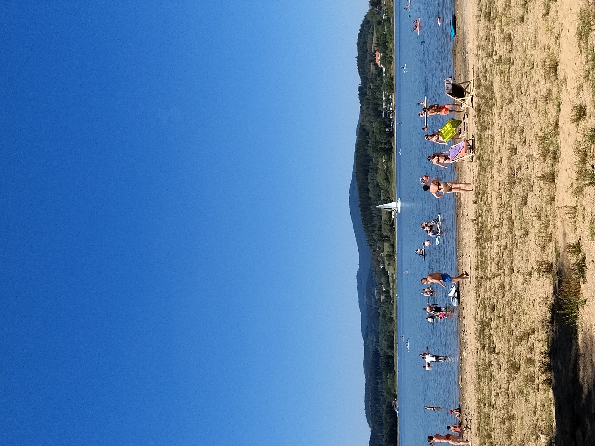 In Radslav, Czechia, a serene lake scene unfolds under a clear blue sky. The foreground is dominated by a sandy beach sparsely populated with dry grass. Several people are visible on the beach. To the left, a woman in a bikini stands facing away from the camera, while a child sits on the sand nearby. Further right, a man in blue shorts walks across the sand. Two lounge chairs, one with a colorful striped cushion and the other a dark canvas, are set up on the beach. A green inflatable raft lies on the sand near the chairs.
The lake itself is calm and blue, with numerous individuals engaged in various water activities. Several people are standing in the shallow water. Scattered across the lake are several paddleboards and a small sailboat. People are seen standing and sitting on the paddleboards, some with visible paddles. Further in the distance, a lush, green treeline forms a backdrop to the lake, with rolling hills and mountains beyond. A few buildings are visible nestled amongst the trees on the far shore. The overall atmosphere is one of peaceful recreation on a warm, sunny day.