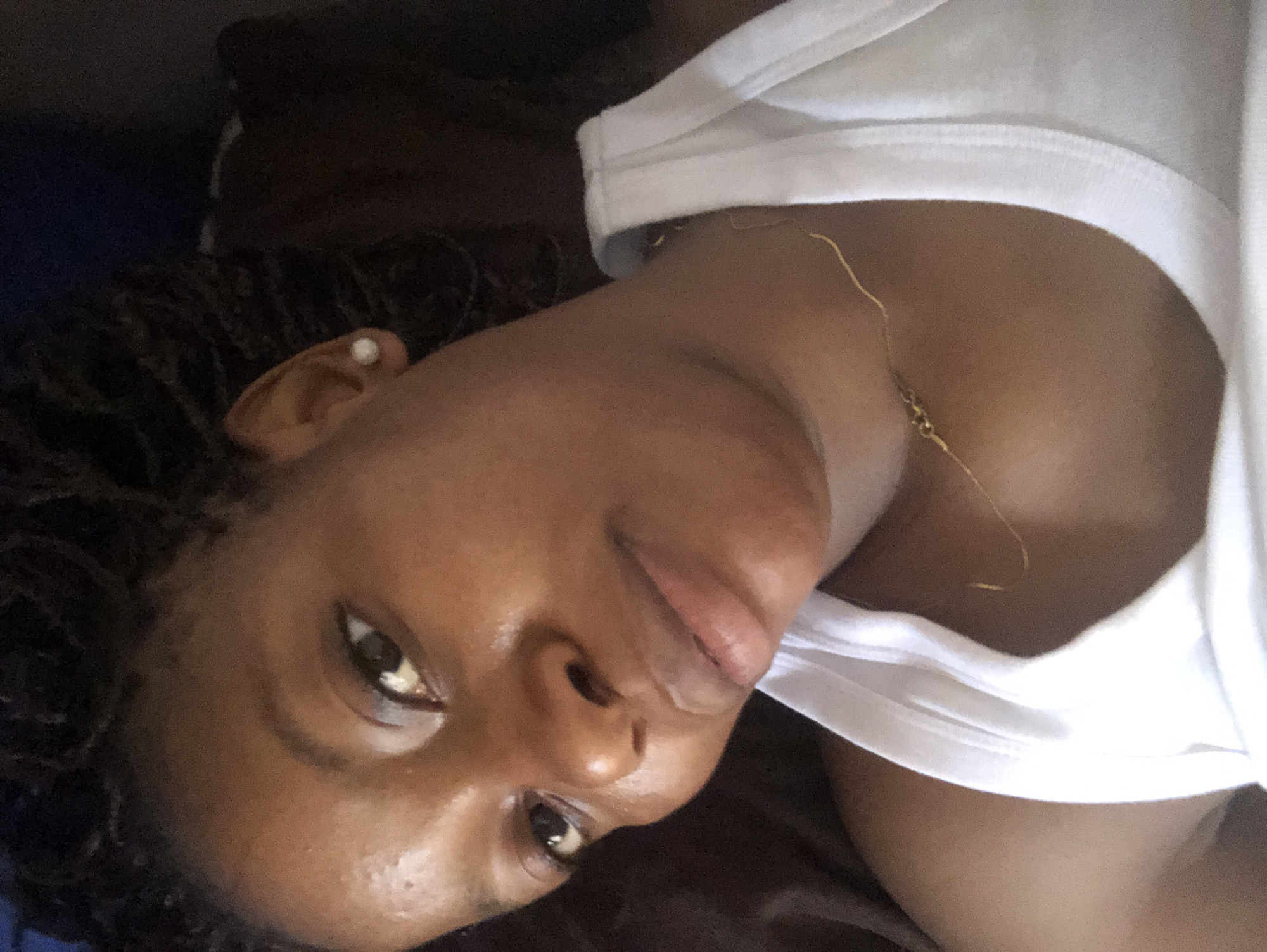 A close-up shot depicts a person with dark skin lying down, wearing a white top and a delicate gold necklace. Their hair is styled in small braids or twists. A single pearl earring is visible in their left ear. The person is looking at the camera with a slight smile. The background is dark and indistinct, suggesting an indoor setting. The location in the city of Kashere, Nigeria cannot be verified from the image.