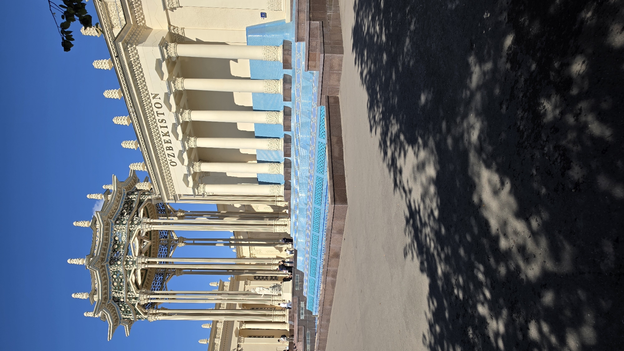 The image shows the pavilion of Uzbekistan at the All-Russian Exhibition Center (VDNKh) in Moscow, Russia. The pavilion, constructed in the Soviet era, features a grand facade with classical columns and an ornate roof. In front of the pavilion is a multi-tiered fountain, decorated with blue tiles and intricate patterns, filled with water. The sky is clear and blue, indicating a sunny day. The ground in front of the fountain is paved with asphalt, and tree shadows are cast across it. Several people are visible near the pavilion and the fountain, suggesting a public space that is actively visited.