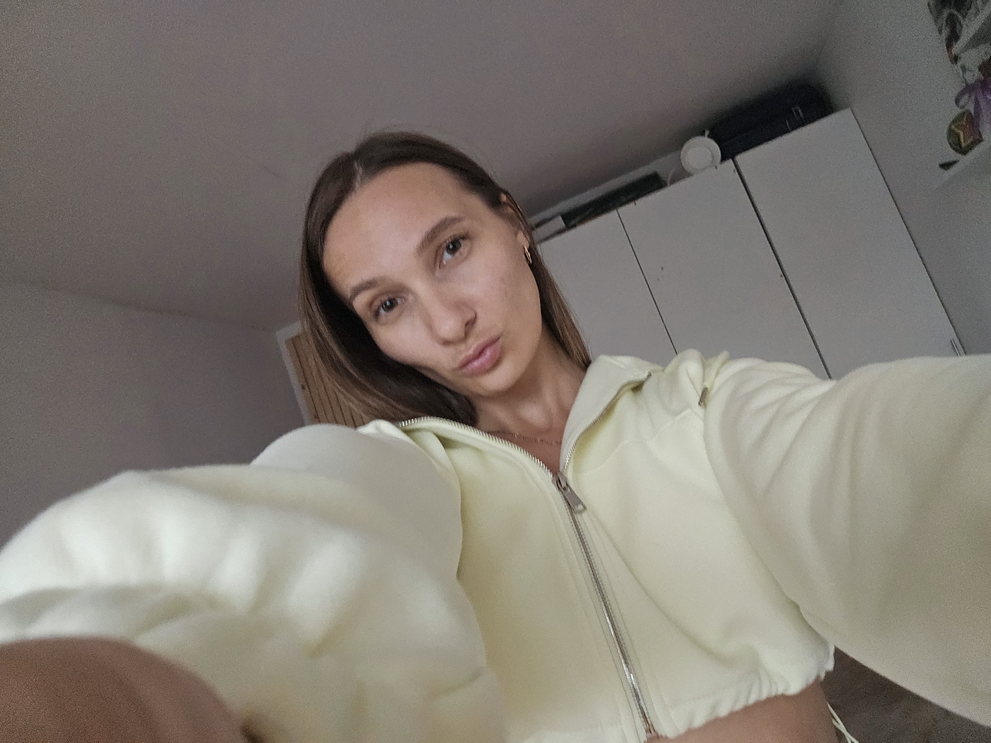 This is a first-person perspective, indoor, medium-shot photograph taken by a young adult female. The subject is positioned in the lower half of the frame, looking directly at the camera with her lips pursed. She has long, brown hair and is wearing a light yellow cropped zip-up jacket. The background consists of plain, light-colored walls and what appears to be a closet or wardrobe. To the right, on top of the wardrobe, a dark suitcase is visible, along with a small, white circular object. The overall lighting is somewhat dim, suggesting an indoor environment. The photograph does not provide any specific visual cues that would indicate the location within the city of Chaikovsky, Russia.