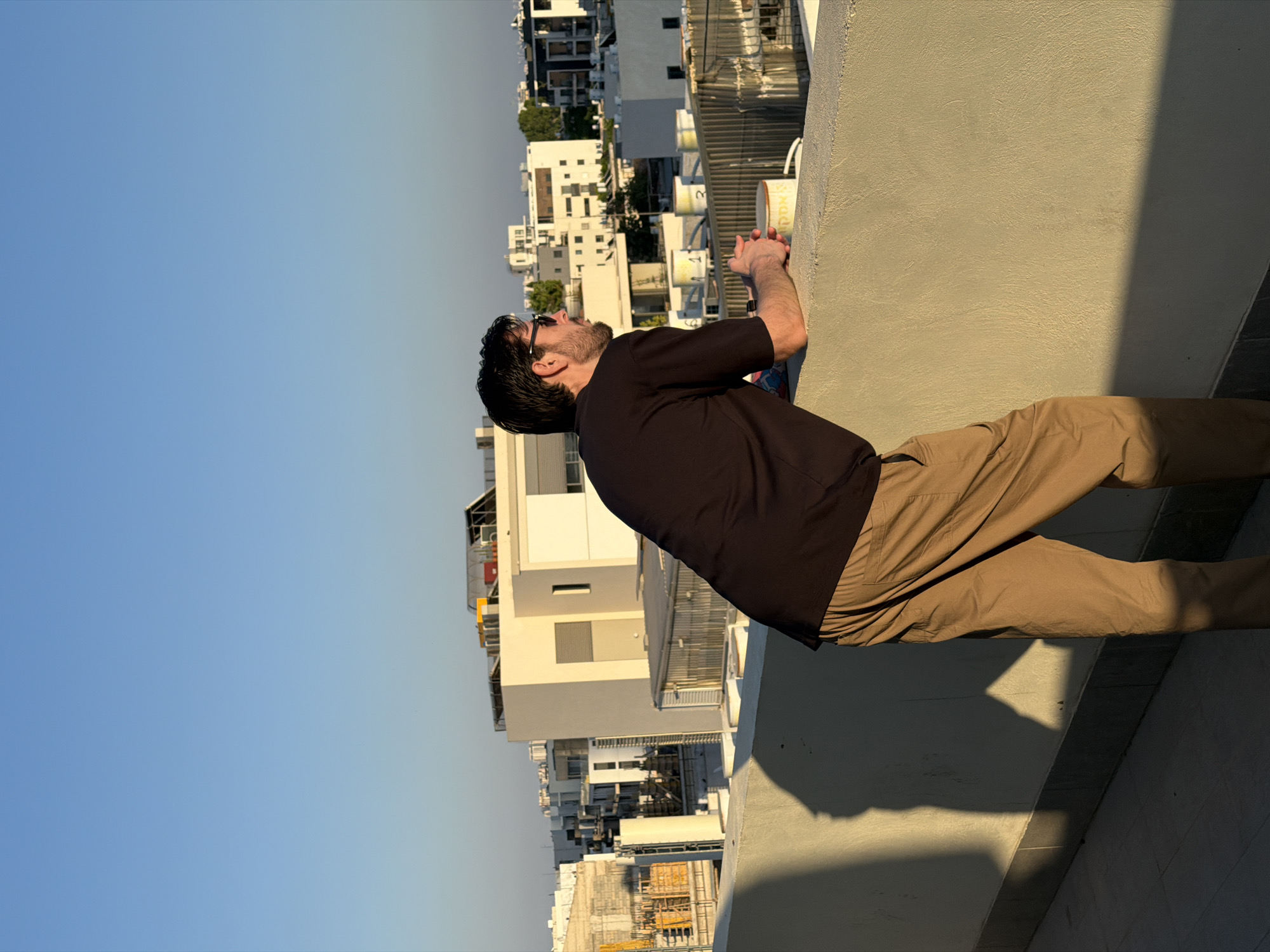 An adult male stands on a rooftop in Tel Aviv, Israel, looking out over the city skyline. He is wearing a black t-shirt and tan pants. His hands are clasped over a low wall, and he is leaning forward, observing the urban landscape. The sun is setting, casting long shadows across the rooftop and bathing the buildings in a warm, golden light. The background shows a mix of modern architecture, including apartment buildings and construction sites. The sky is clear and blue, with a gradient towards orange near the horizon.