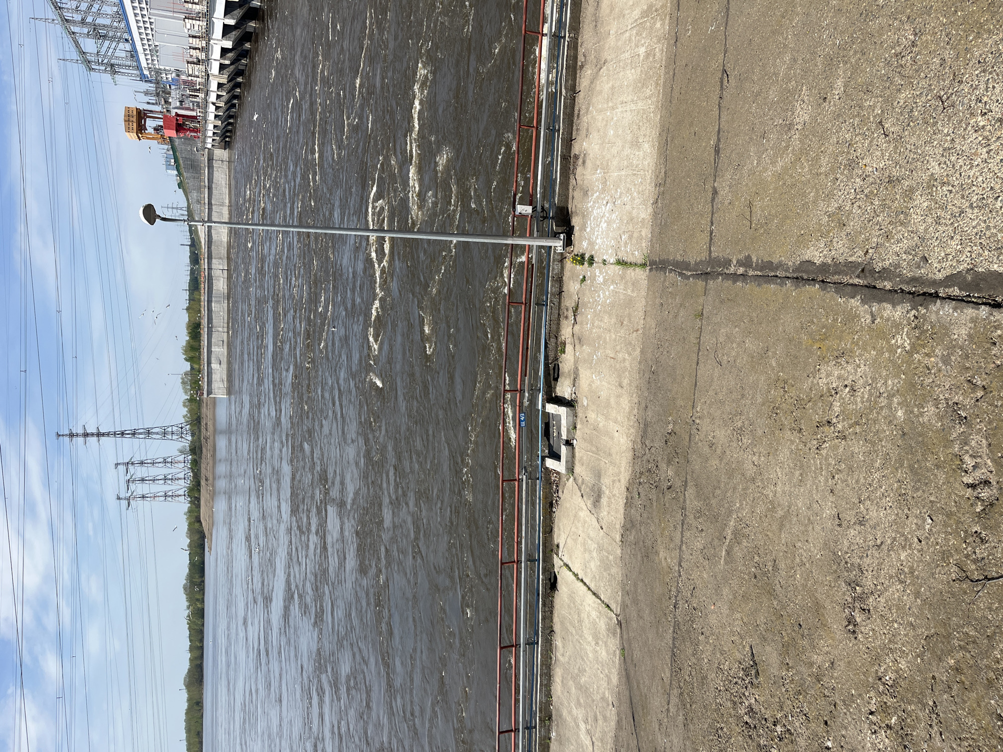 The image shows a section of the dam on the Kama River in Chaikovsky, Russia. The water is turbulent and flowing rapidly. A concrete wall and a metal railing separate the water from a concrete walkway. A lamppost stands near the railing. In the background, there is a hydro-electric power station and power lines. The sky is partly cloudy.