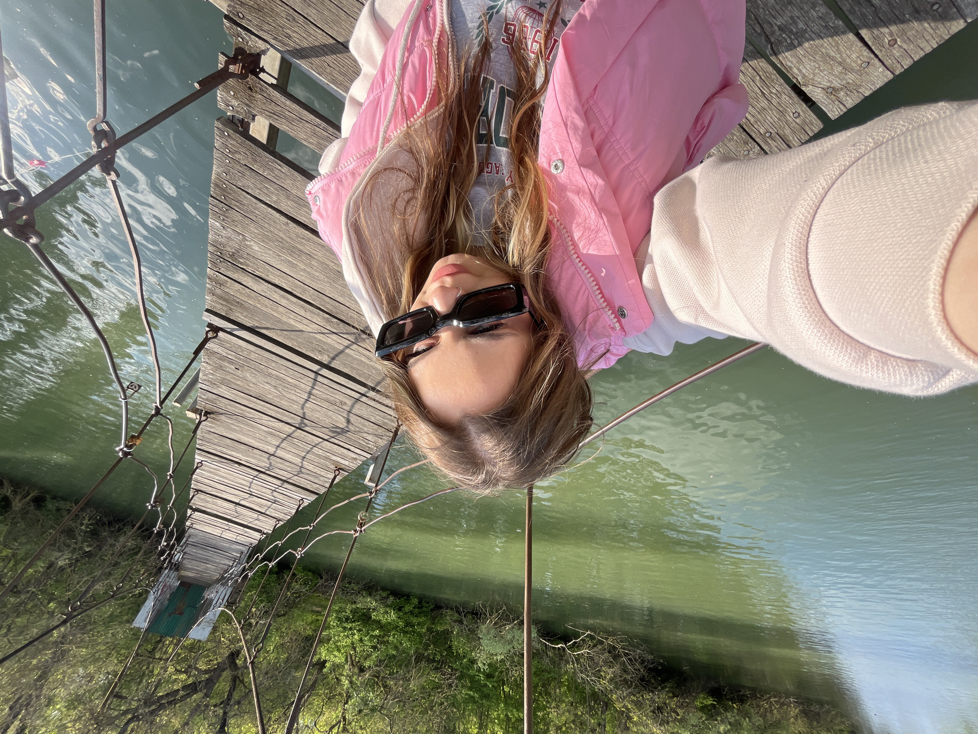 A woman wearing sunglasses, a gray sweatshirt, and a pink jacket is lying on her back on a wooden suspension bridge. The bridge spans a body of water, with trees visible on the opposite bank. The photograph is taken at an oblique angle looking directly at the woman. The location is likely Krasnodar, Russia based on visual features of the bridge.