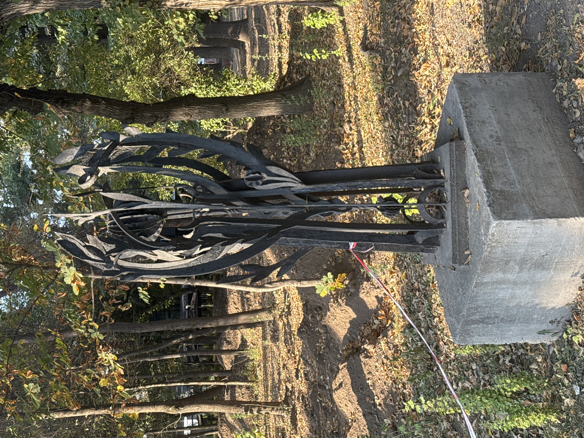 A black metal sculpture resembling stylized trees or flames stands on a concrete base in a park-like setting. The sculpture is dark and abstract, with thin, branching elements. The concrete base is rough and weathered, with some dirt and leaves accumulated on it. Around the sculpture, the ground is covered in fallen leaves, indicating an autumn season. There are trees with green and yellowing leaves in the background, suggesting a temperate climate. A red and white striped tape runs across the foreground, partially obscuring the base of the sculpture, possibly indicating a restricted area. The lighting suggests it is daytime, with sunlight casting shadows. The overall scene appears to be an outdoor public space, likely a park or garden.