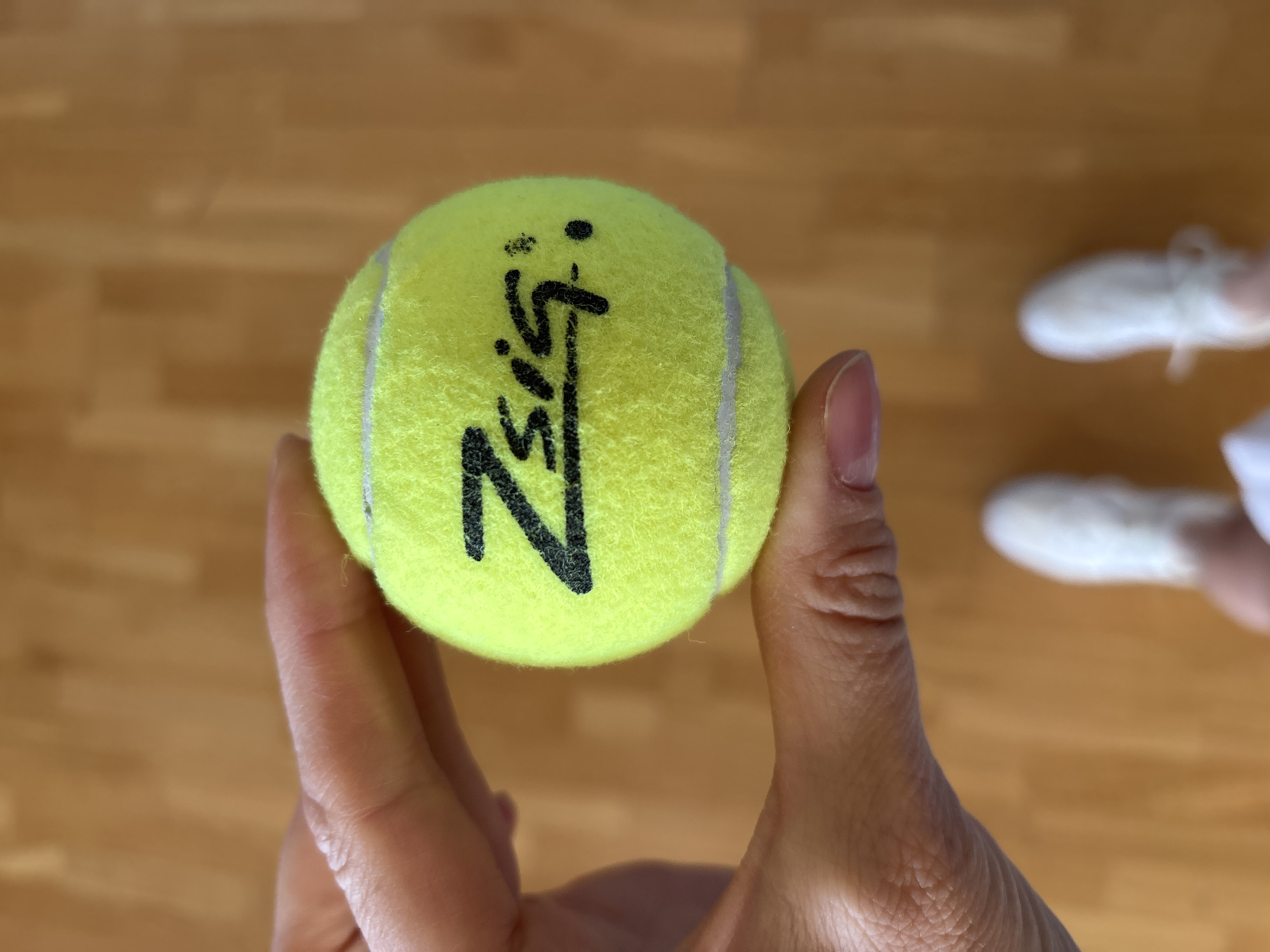 An overhead shot captures a person's hand holding a bright yellow-green tennis ball in an indoor setting, presumably in Chaikovsky, Russia. The hand is positioned centrally, holding the fuzzy tennis ball towards the viewer. The background features a light brown wooden floor, blurred due to a shallow depth of field, with two feet wearing white athletic shoes or sneakers also visible and out of focus.
The primary activity is the presentation of the tennis ball. The ball itself is a standard felted tennis ball with visible white seam lines. A prominent, stylized black marking or signature is inscribed on the ball. This writing appears to spell "Zizi" or "Ziss," featuring a distinct "Z" at the beginning, followed by an "i" or similar letter, then a character that could be an "s" or another "i," and concluding with a stylized mark. There are also two small black dots positioned above the middle section of the signature. The person's hand has visible skin texture and neatly trimmed fingernails. The white shoes in the background are casual or athletic, featuring white laces, further suggesting a casual or sports-related environment. The lighting suggests an interior space, likely with natural or artificial ambient light.