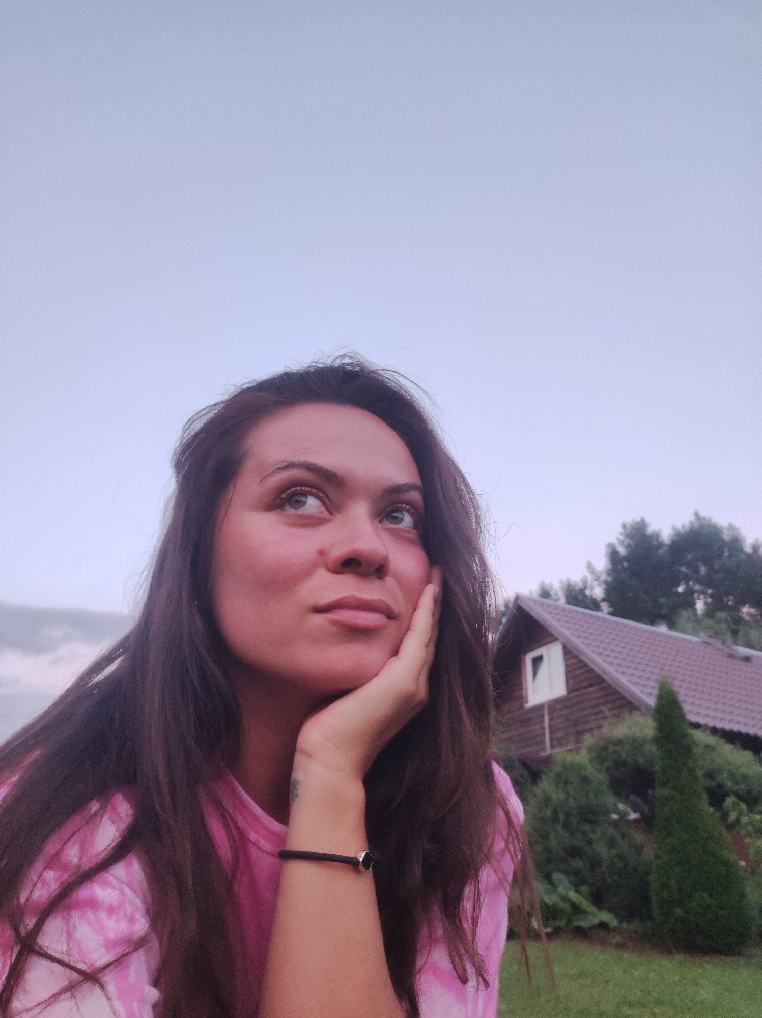A young woman with long, dark brown hair is pictured in a close-up, outdoor shot. She is looking upwards and to her right, with her right hand resting on her chin and her cheek. Her left arm is bent, with a black bracelet on her wrist. She is wearing a pink tie-dye t-shirt. In the background, a wooden house with a dark roof and a white-trimmed window is visible, surrounded by green trees and grass. The sky is a pale, overcast blue. The scene appears to be set in a rural or suburban area, possibly in or around the city of Fokino, Russia, given the architectural style of the house and the surrounding vegetation.