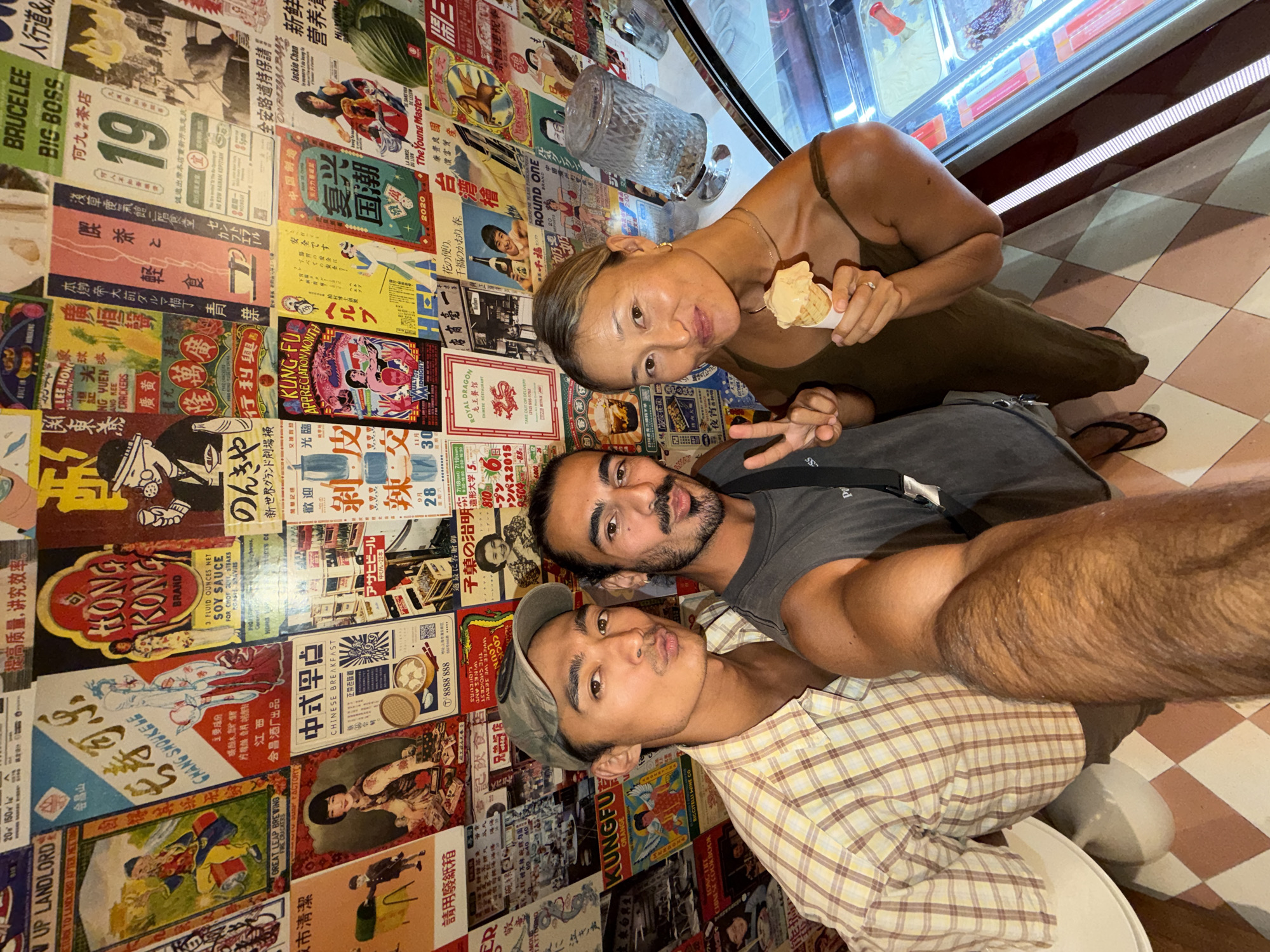Three people are taking a selfie in front of a wall covered in posters. A woman is holding an ice cream cone. A man behind her is making a peace sign. There is an ice cream display case next to the woman and a glass jar on top of it. The floor is tiled. The image was taken in Mengwi, Indonesia.