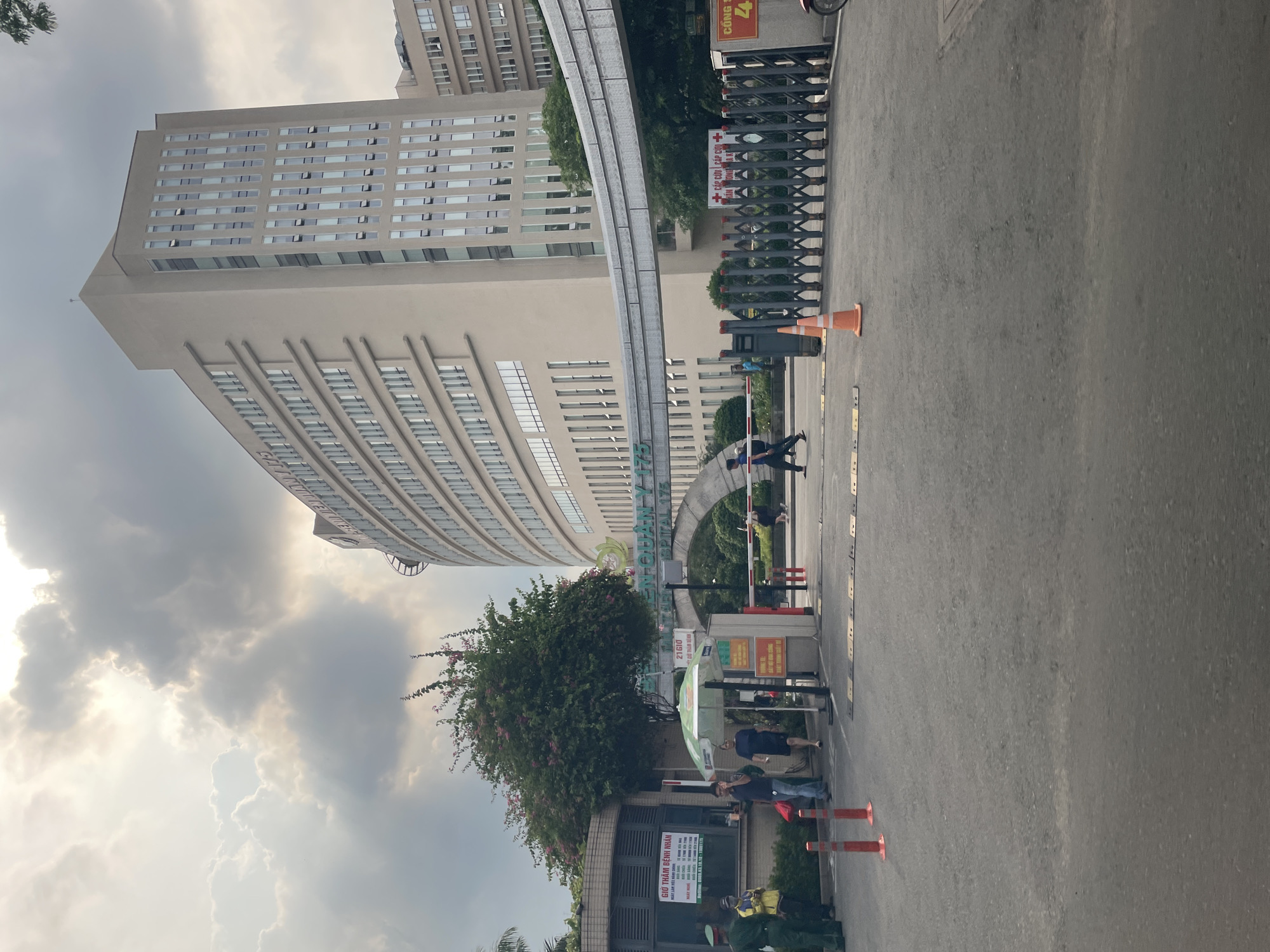 The image captures the entrance to the Thủ Đức District Hospital in Thủ Đức City, Vietnam, on a cloudy day. A large, modern, beige-colored hospital building dominates the background, with numerous windows and several floors. A curved pedestrian overpass, also beige, spans across the entrance area.
In the foreground, a paved road leads into the hospital grounds, with orange traffic cones and speed bumps visible. A dark metal folding gate is partially open, controlling access to the hospital. To the left, a guard booth with a sign is visible, and two individuals in green uniforms, presumably security personnel, stand near it. Several other people are seen walking in the entrance area, some under a green and white umbrella.
A prominent sign arching over the entrance reads "BỆNH VIỆN QUẬN 175" (Hospital District 175) in green lettering, with "HOSPITAL 175" below it. Another sign near the gate displays "CẤP CỨU" (Emergency) with a red cross symbol. A large tree with pink flowers is situated to the left of the entrance arch. The sky is filled with dramatic, heavy clouds, suggesting overcast or impending rain.