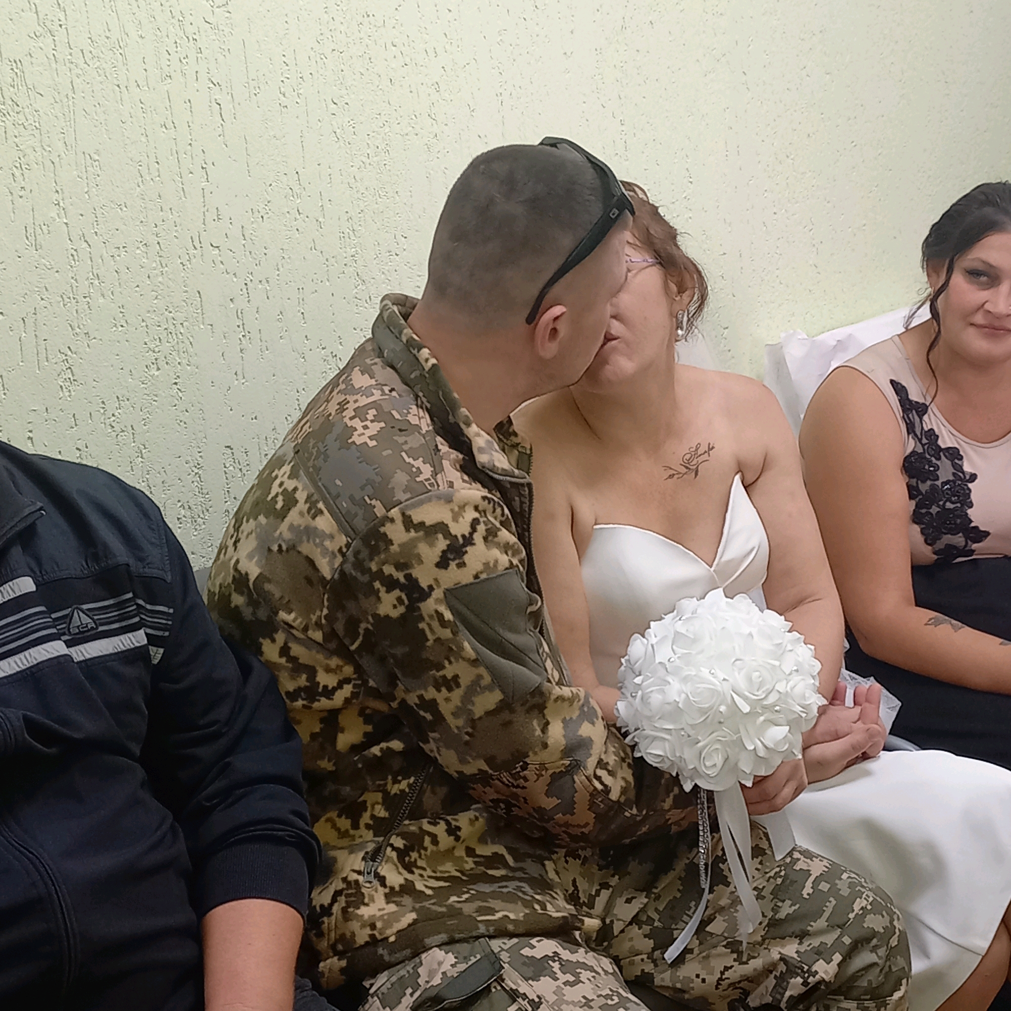 In Pervozvanivka, Ukraine, a newly married couple is captured in an intimate moment, kissing, with a woman in a beige dress with black floral applique observing from the right and another person partially visible on the left. The groom, wearing a camouflage military uniform, holds a bouquet of white roses. The bride is in a white strapless wedding dress. The background is a textured, pale yellow wall.