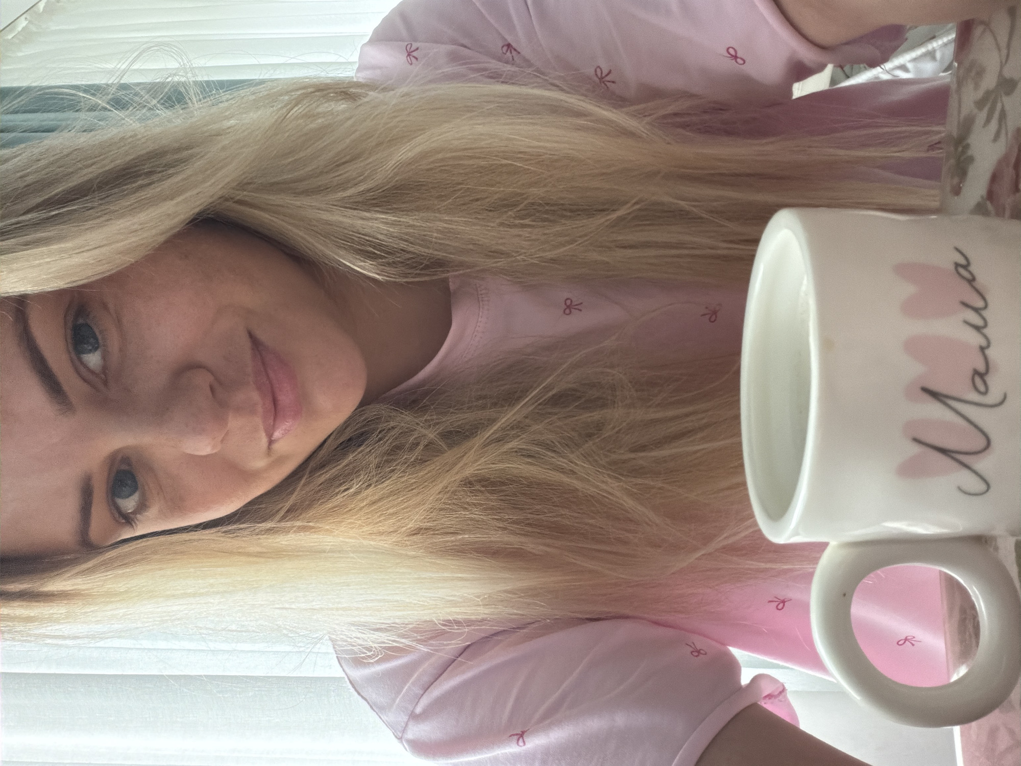 The image shows a close-up of a blonde woman with blue eyes and fair skin, wearing a pink t-shirt with small red bows. She is smiling slightly and looking towards the camera. In the foreground, a white mug with the word "Mama" and two pink hearts is visible. The background consists of a white curtain. This scene is not located in Votkinsk, Russia.