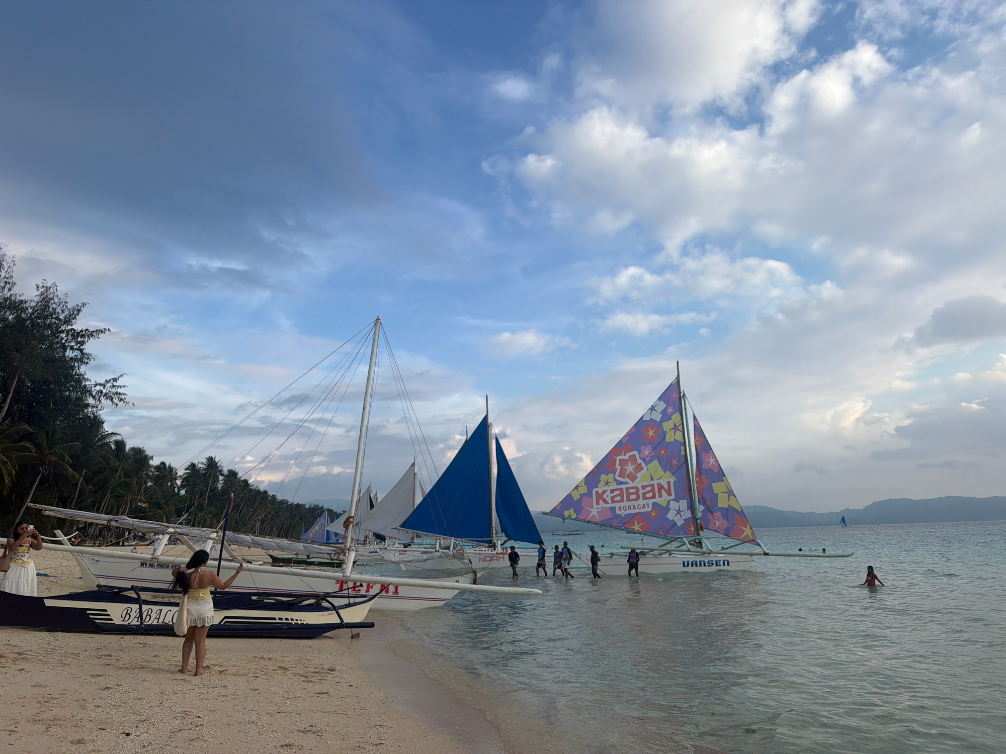 This image captures a picturesque scene at White Beach, Boracay, in the city of Malay, Philippines. The foreground features a sandy beach with several traditional Filipino outrigger boats, known as paraws, lined up along the shore. Two women are visible on the beach, one in the foreground posing, and another further back taking a photo. In the water, several paraws are sailing, with their colorful sails adding vibrancy to the scene. Some people are wading in the shallow water near the boats, while others are seen swimming further out. The background displays a tranquil sea under a sky filled with scattered clouds, with a faint outline of islands in the distance. The overall atmosphere is serene and inviting, typical of a tropical beach destination.