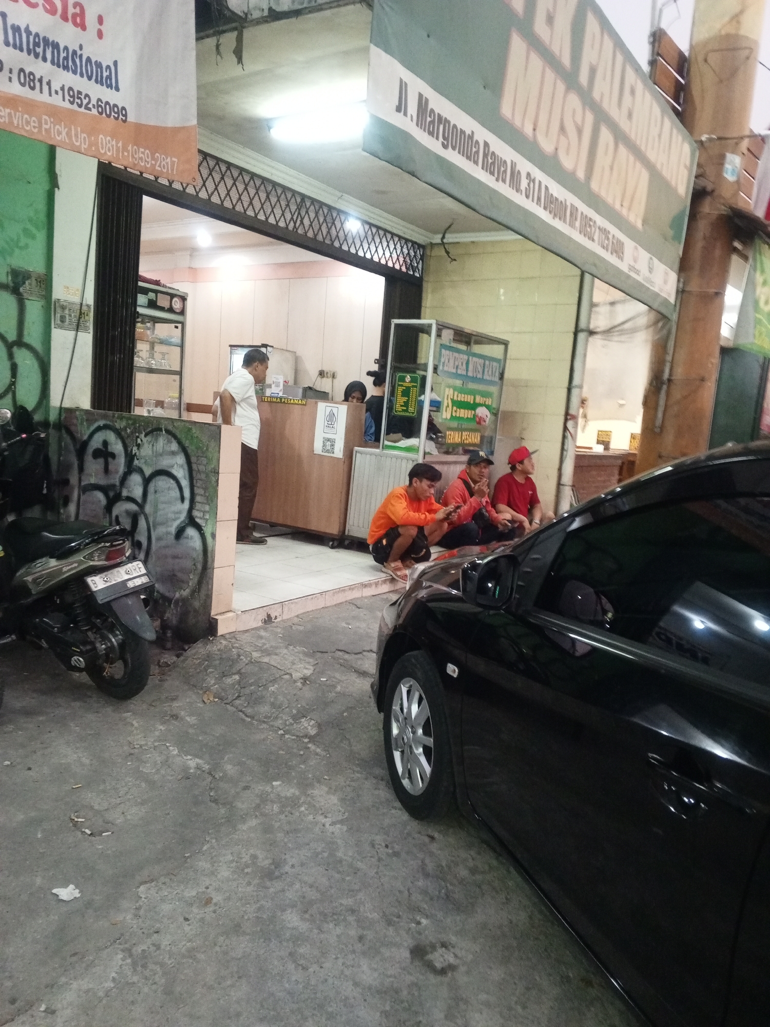 In Depok, Indonesia, in front of a restaurant called "PEMPEK PALEMBANG MUSI RAYA" located on Jl. Margonda Raya No. 31A, a group of people are gathered. A black Toyota car is parked in the foreground, its side and front wheel visible. To the left of the car, a motorcycle is parked against a graffiti-covered wall. Inside the restaurant's open doorway, a man in a white shirt stands near a wooden counter. Three young men are seated on the ground to the right, one in an orange shirt, another in a red jacket, and the third in a red cap. They appear to be casually interacting or waiting. Signs are visible above the entrance, one with Indonesian text and another indicating the restaurant's name. The ground is paved with concrete and appears worn.
