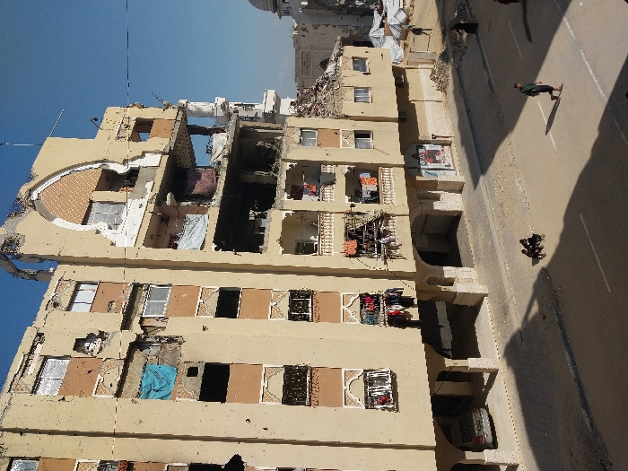 This image captures a multi-story, beige-colored building in Khan Yunis, Palestinian Territory, exhibiting extensive structural damage under a clear blue sky. The upper right portion of the building's facade and roof are missing, revealing interior floors and a pointed arch architectural detail. Numerous small, dark impact marks, consistent with bullet or shrapnel damage, are visible across the building's exterior walls. Many windows are broken or entirely absent. Various pieces of clothing, including a light blue sheet and assorted garments, hang to dry from multiple balconies on the building's front.
At street level, the paved road is marked by strong shadows cast from the left, indicating bright sunlight. Two small children are seated on the street in the left-center. An adult male, wearing a green top and dark pants, walks from right to left across the street, appearing to be barefoot or in minimal footwear. Another individual in dark clothing is visible further down the street to the right. In the background, beyond the immediate building, widespread destruction is evident, with piles of rubble and debris. A white dome structure, possibly a mosque, and a white minaret are partially visible in the distant background, amidst additional damaged structures and temporary white tarp shelters. A silver sedan is parked beneath an arched ground-floor area of the main damaged building.