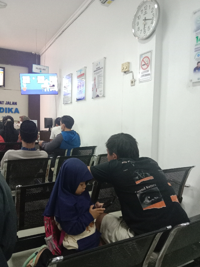 The image displays an indoor waiting area within a public service facility or clinic in Gladaksari, Indonesia. The environment features white walls and multiple rows of black, perforated metal-frame waiting chairs. An analog clock on the wall indicates approximately 9:07. Below the clock, a "No Smoking" sign, with Indonesian text "DILARANG MEROKOK DI AREA INI," is visible alongside beige electrical outlets. Several informational posters in clear frames are mounted on the wall. To the left, a flat-screen television displays animated graphics and text, likely public service information or a queue system, with a sign reading "POJOK JKN" suspended above it.
In the foreground, a young child, wearing a dark blue hijab and a light-colored shirt, is seated on a chair, actively operating a dark-colored smartphone. A pink bag is situated beside her. To her immediate right, an adult male is seated, wearing a dark t-shirt featuring "Sound Nature" text and graphics, and light-colored shorts. He is oriented towards the child with his back partially to the camera.
In the mid-ground and background, several other individuals occupy the waiting chairs. One person wears a light grey long-sleeved shirt and a black cap or kufi, facing away from the camera. Another person wears a blue hooded jacket and a white face mask. Multiple other individuals are partially visible or out of focus in the seating area. A desk with a computer monitor is discernable in the left background.