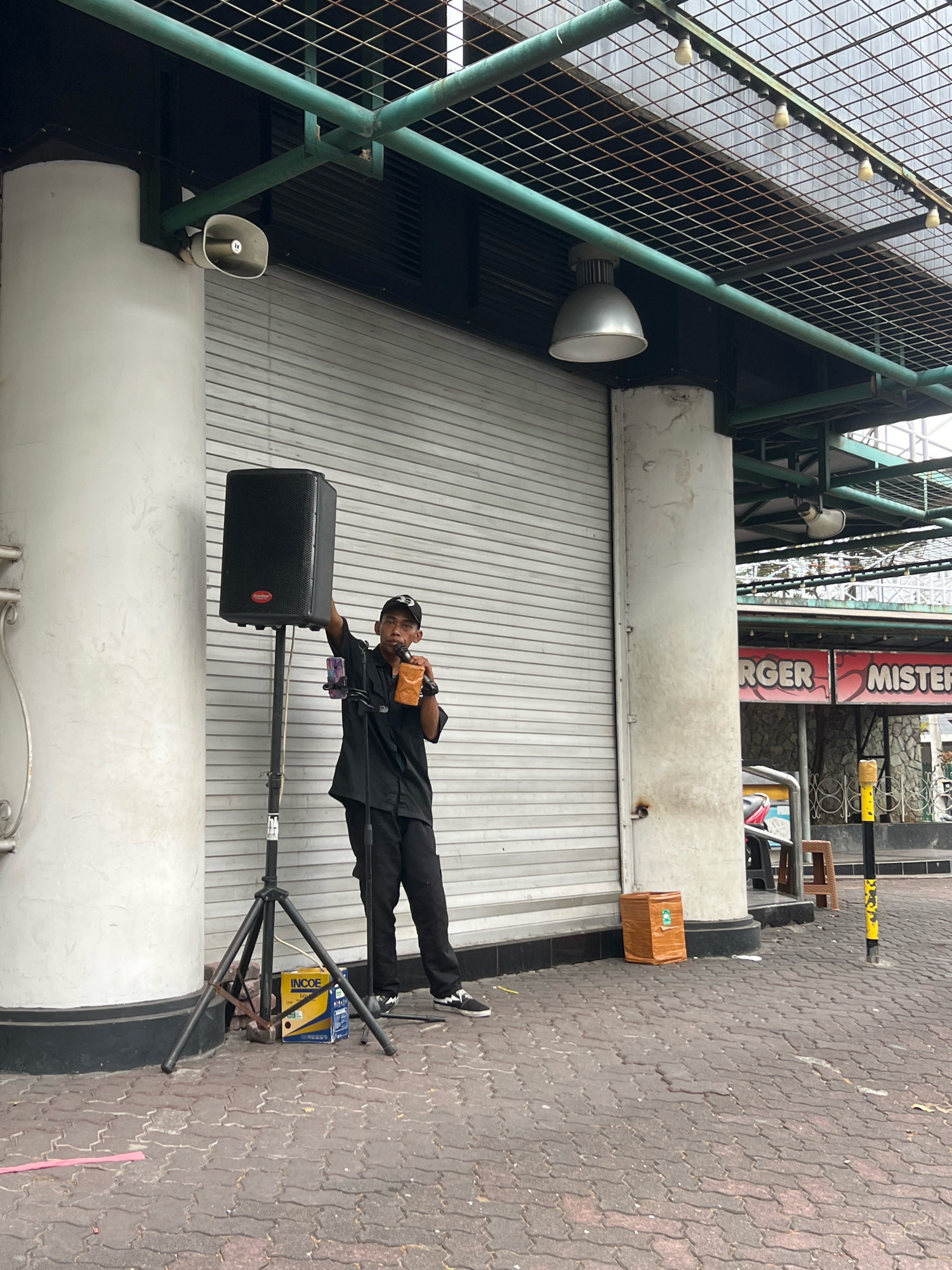 A person wearing a black shirt, black pants, and a black baseball cap stands under a green metal awning in front of a closed corrugated metal storefront. The person is holding a drink in their right hand and a microphone in their left hand, appearing to speak into it. A black speaker is mounted on a tripod to the person's left. A smartphone is attached to the tripod, facing the person. A yellow and blue box is on the ground near the tripod. In the background, a sign reads "RGER MISTER". The ground is paved with red interlocking bricks. The scene appears to be outdoors in an urban environment.