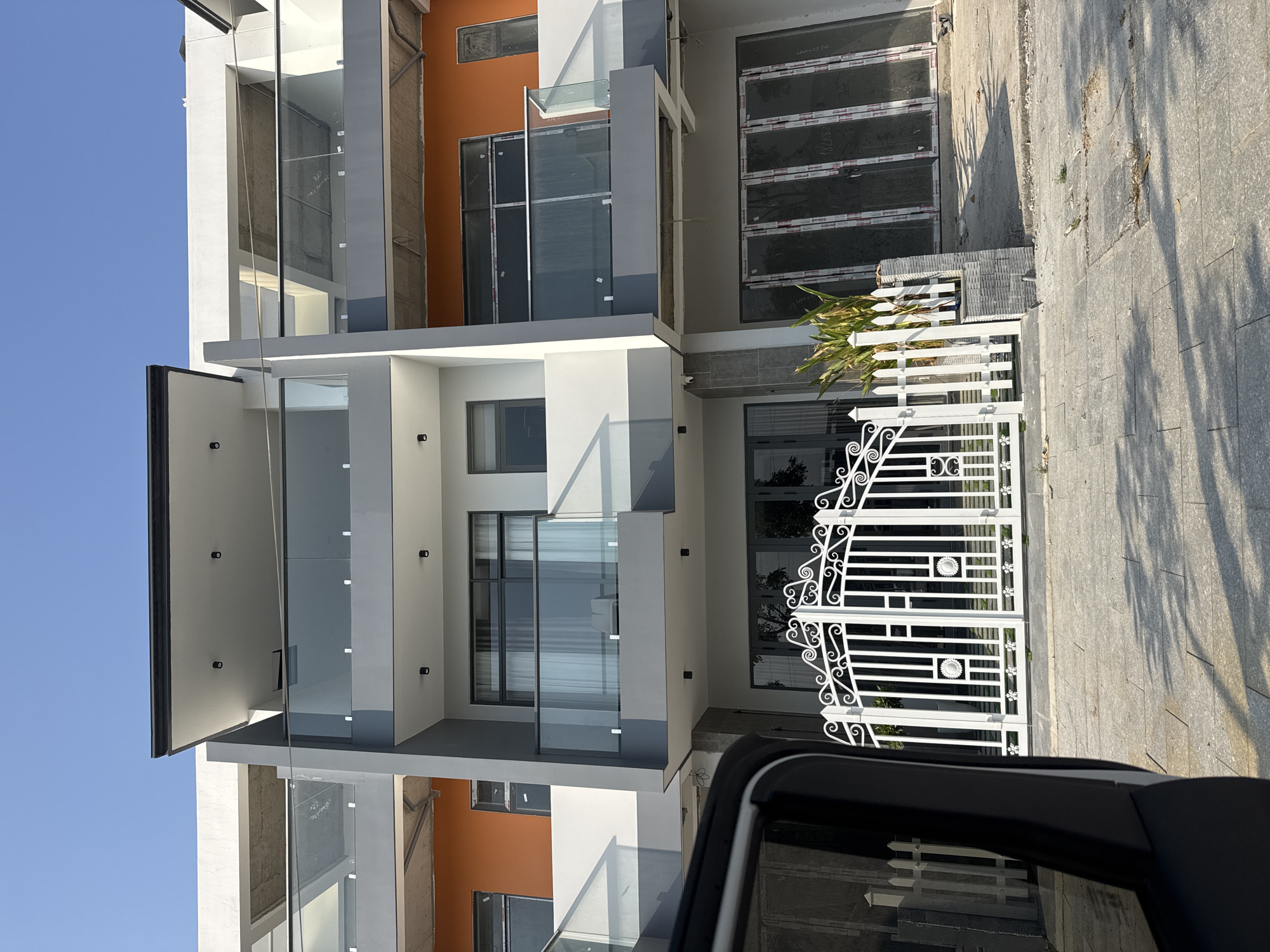 Here is a description of the image:
The image captures a modern building with a combination of white, gray, and orange colors. The architecture is characterized by multiple levels and a glass-panel balcony design. A white, ornate gate stands at the front of the property, partially visible behind the edge of a black vehicle. The surrounding area appears to be a paved street with shadows cast by sunlight, suggesting an urban or suburban location. The scene is devoid of people.