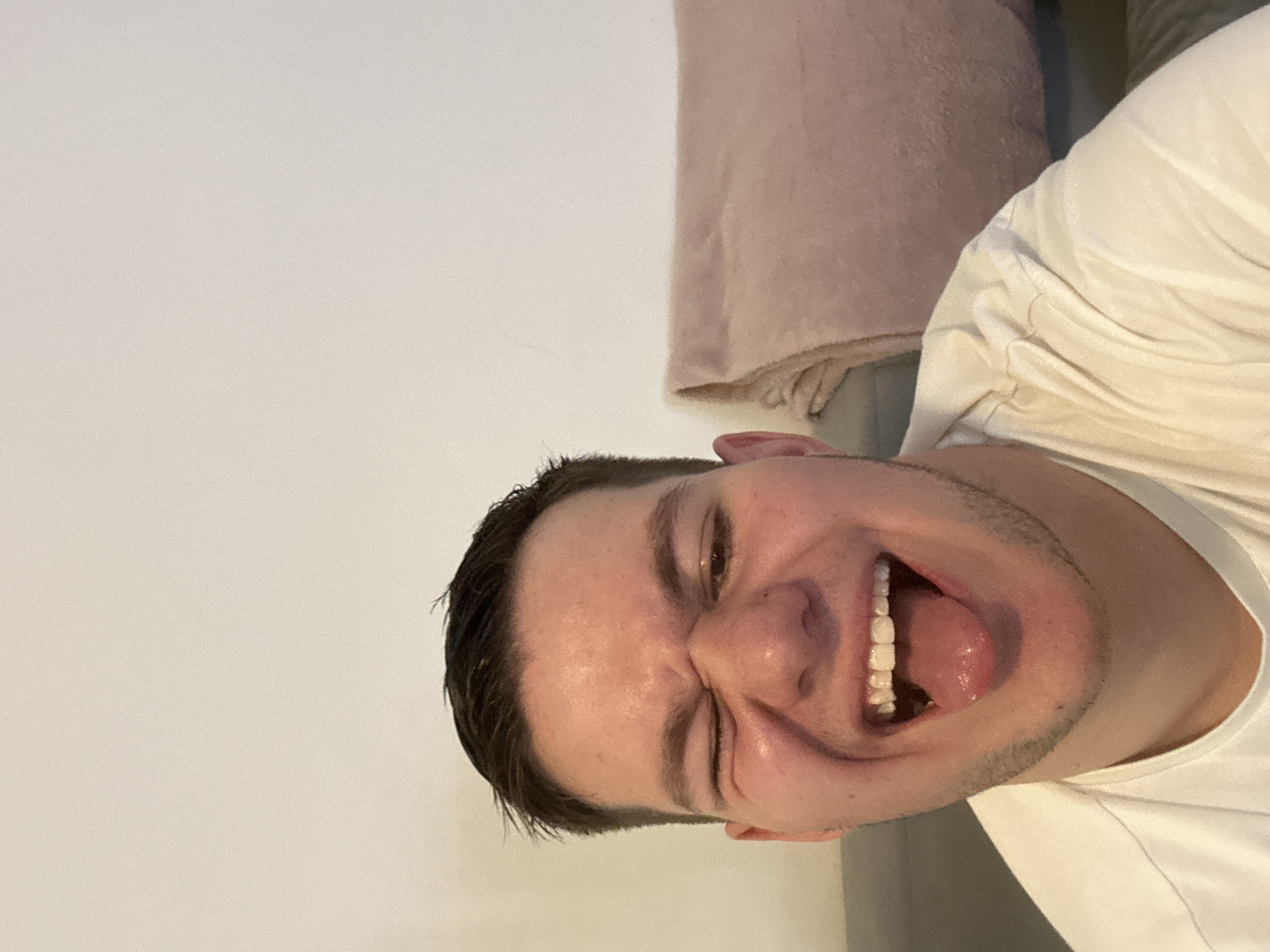 A close-up, low-angle, rotated image shows a fair-skinned male with dark hair and stubble, wearing a white shirt. He is grinning widely with his tongue sticking out. Behind him, there is a cream-colored wall and a pink pillow. It is not possible to determine the location in the city of Sofia, Bulgaria from this image.