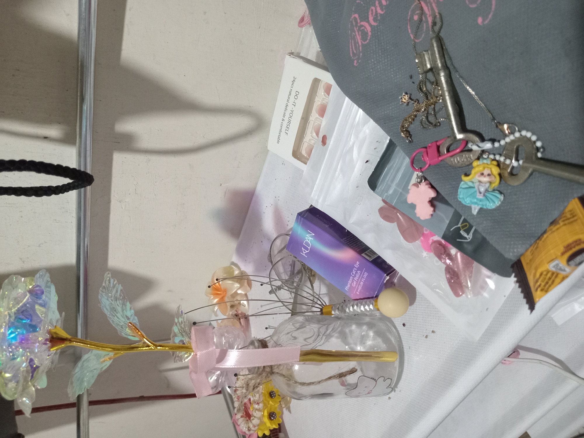 The image depicts a shelf in Claket, Indonesia, displaying a variety of personal items. In the foreground, a collection of keys with charms hangs from a grey fabric surface. Among the charms are a small doll and a pink cloud-shaped pendant. To the left, a clear glass bottle with a pink ribbon and a golden stem is visible, containing a decorative flower. Beside it, a purple box labeled "KUDAN Phantom Cat's Eye Gel Polish" and a box of artificial fingernails are present. In the background, a black rope hangs from a metal rod. The overall scene suggests a personal vanity or collection area.