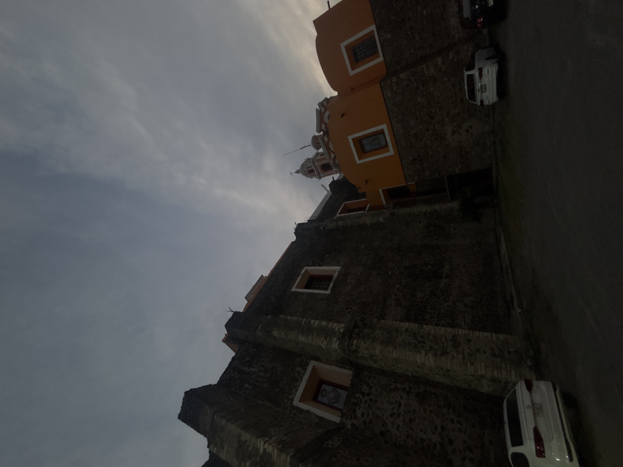 The image depicts a low-angle shot of a building and the sky above. The building appears to be old and made of stone, with an orange addition on the upper part. The building’s windows are visible, and several cars are parked on the adjacent road. The sky is overcast with a muted color palette. The location is San Andrés Cholula, Mexico.