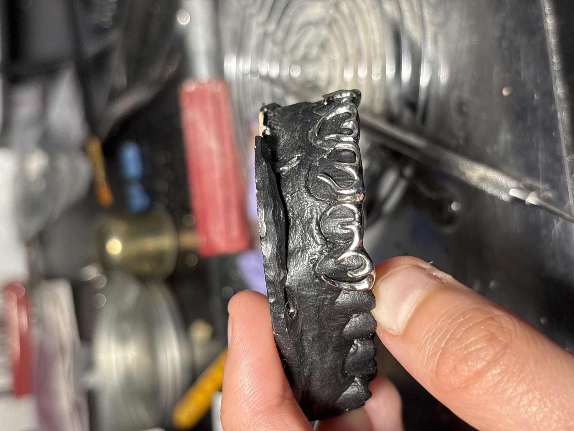 A close-up image shows a person holding a black, intricately designed object. The object appears to be made of a dark, possibly stone-like material, with silver-colored, decorative elements embedded within it. The person's fingers are visible, gripping the object. The background is cluttered and out of focus, showing various tools and metallic surfaces, indicating a workshop or studio environment. There is no information available in the image to indicate that it was taken in Sofia, Bulgaria.
