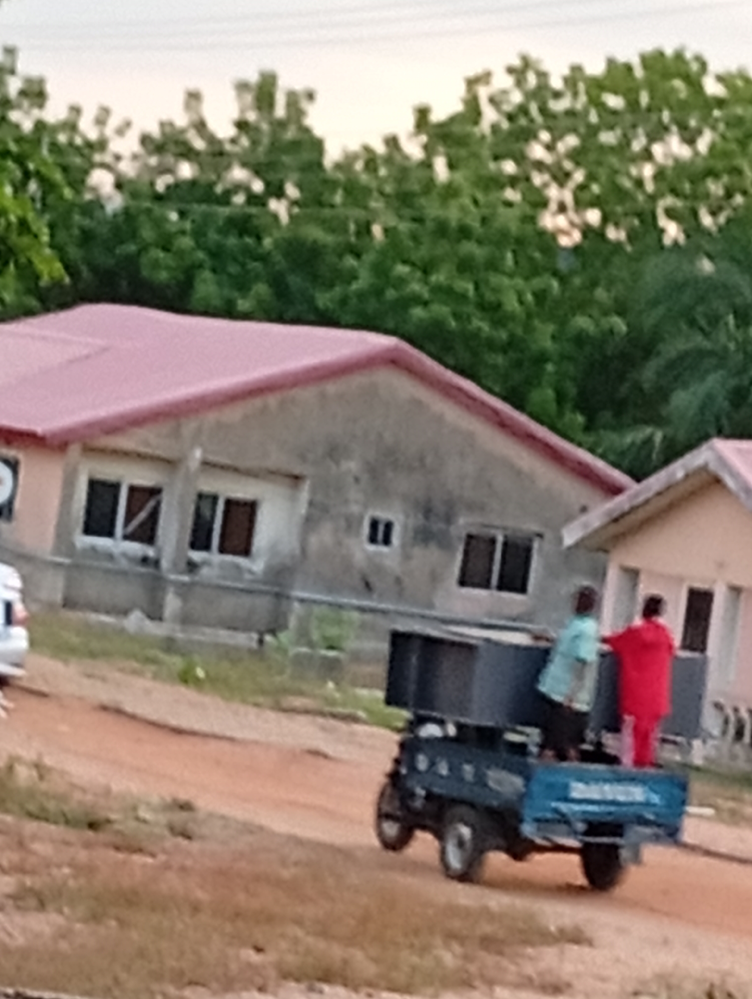 A slightly out-of-focus, medium-shot depicts two people on the back of a blue, three-wheeled vehicle on a dirt road in front of buildings. The first building is gray with a red roof and four windows. The second building is peach-colored, also with a red roof. Trees are visible in the background. The first person is wearing a light blue shirt and the second is wearing a red robe-like garment. The vehicle is loaded with dark objects. The location is Unknown, Unknown.