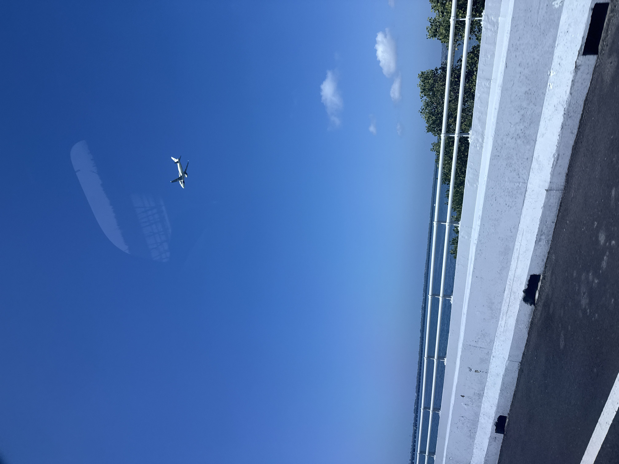 A white airplane is visible in the clear blue sky, flying high above a body of water. In the foreground, a white concrete barrier with metal railings separates the water from a road. The scene appears to be a bridge overlooking the ocean.