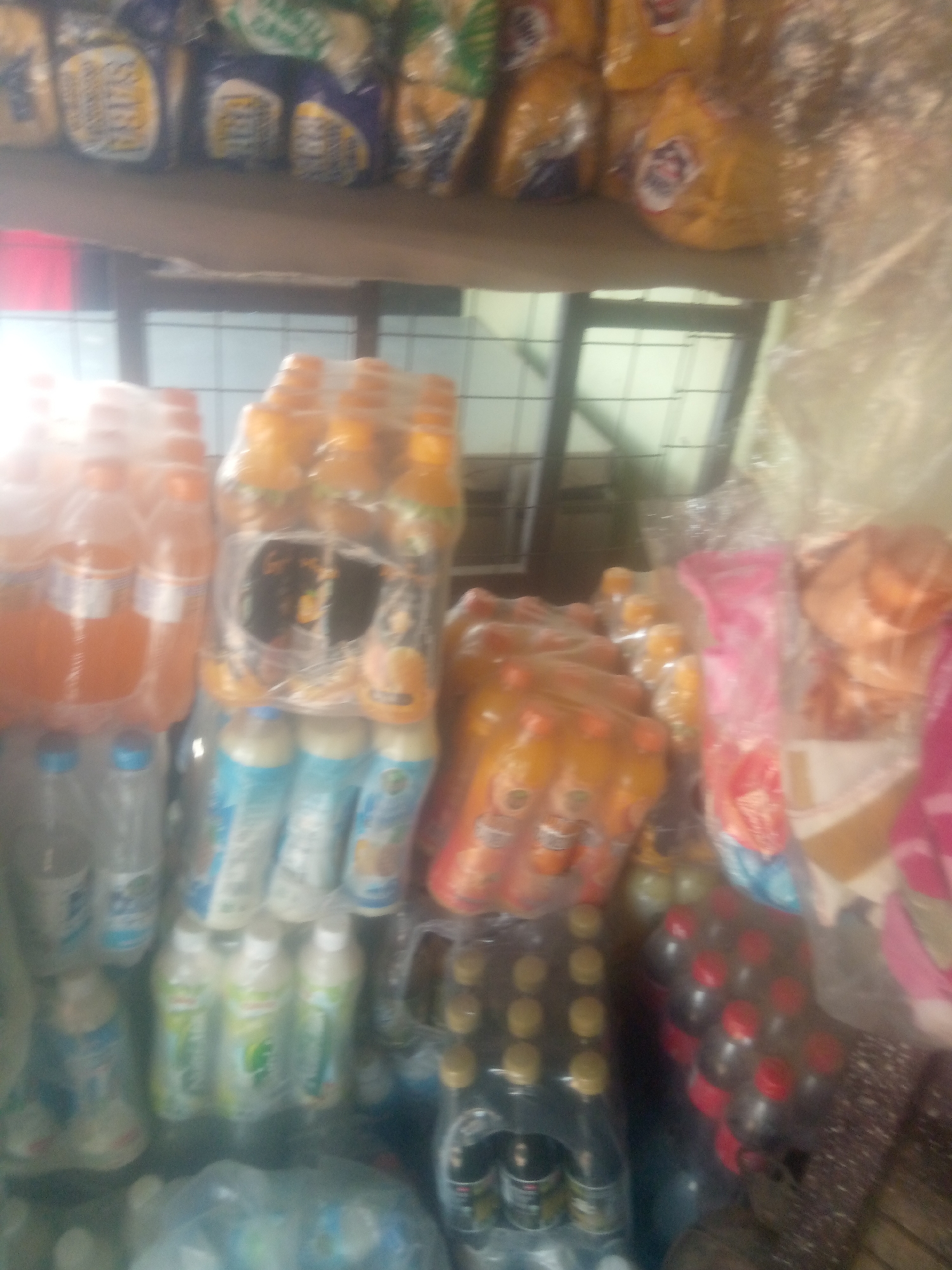 The image is a blurry, overhead shot of a shelf stocked with various beverages, likely in a store in Jalingo, Nigeria. The shelf appears to be made of wood or a similar material. On the top shelf, there are several bags of what appear to be snacks or packaged goods, with some yellow and purple packaging visible. Below this, on a lower shelf, a collection of plastic bottles filled with orange-colored liquid is arranged in rows, some wrapped in plastic. To the right of these, more bottles containing a similar orange liquid are visible, along with some bottles of lighter-colored beverages. Further to the right, there are stacks of dark-colored bottles with red caps, also likely beverages. In the foreground, there are more bottles of clear or light-colored liquid, and some items wrapped in plastic. The overall impression is one of a retail setting with a variety of drinks and packaged goods on display. The lighting is somewhat dim and appears to be artificial.