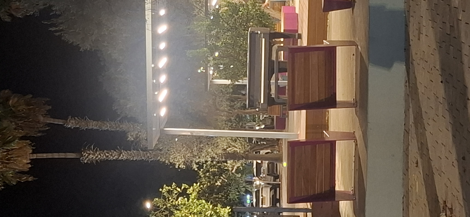The image depicts an outdoor scene at night, possibly a park or public square in Yerocham, Israel. In the foreground, two wooden benches with metal frames face away from the viewer, positioned on a paved surface. Behind the benches, a raised wooden platform serves as a stage. A grand piano sits on this stage, illuminated by overhead string lights. Various trees and foliage surround the stage area, with palm trees visible in the background. A light-colored building or structure with large windows is partially visible on the left, and two cars are parked on a road in the middle ground. The overall ambiance is that of a performance or gathering space set up for an evening event.