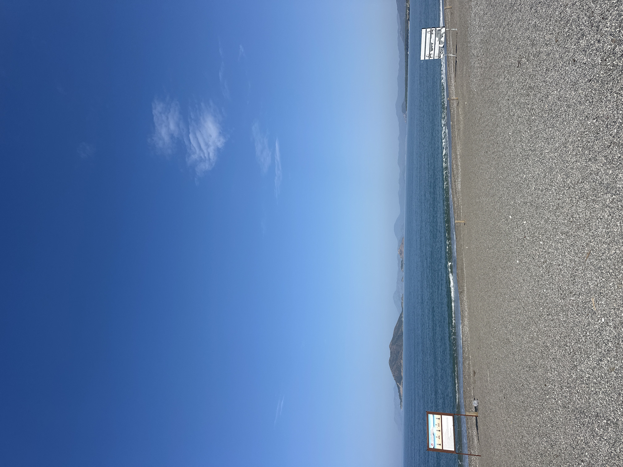 The image depicts a beach scene in Fethiye, Turkey, under a clear, blue sky with a few scattered clouds. The beach is covered in gray pebbles and stretches to the right of the frame. A sign with information is visible near the water's edge. The sea is blue with small waves gently washing ashore. In the distance, mountainous landmasses are visible, adding depth to the landscape. The composition is vertical, with the sky dominating the left side.