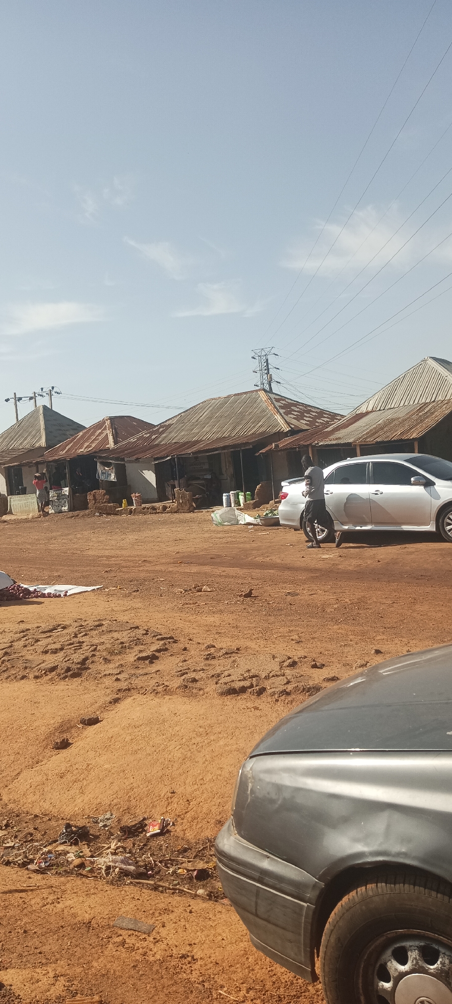 This outdoor scene captures a bustling roadside or market area under a bright, clear sky. The foreground is dominated by reddish-brown, unpaved earth, showing signs of vehicle tracks and uneven patches.
In the middle ground, a row of single-story structures with weathered, corrugated metal roofs line what appears to be a dirt road. These buildings function as shops or stalls, with various goods displayed. On one of the stalls, a sign is partially visible, appearing to read "JINGOTE" or similar.
Several people are present, indicating ongoing activity. On the left, a person in a red top stands near a stall. In the center-right, a man in a dark grey shirt and pants walks alongside a silver sedan, which is parked facing right. Near the stalls, sacks of goods and a white sheet with red produce (possibly onions) are laid out on the ground. A basin containing green items is also visible near the silver car.
The front right portion of a dark grey car, with its wheel visible in the bottom right, occupies part of the immediate foreground. Litter is scattered on the ground near this vehicle. Overhead, numerous power lines crisscross the sky, and a large metal electricity pylon stands prominently in the background.
The time of day appears to be mid-day or early afternoon, given the bright, direct sunlight and lack of long shadows. The setting suggests a semi-urban or rural environment, characterized by the unpaved ground, simple architecture, and visible market activities.