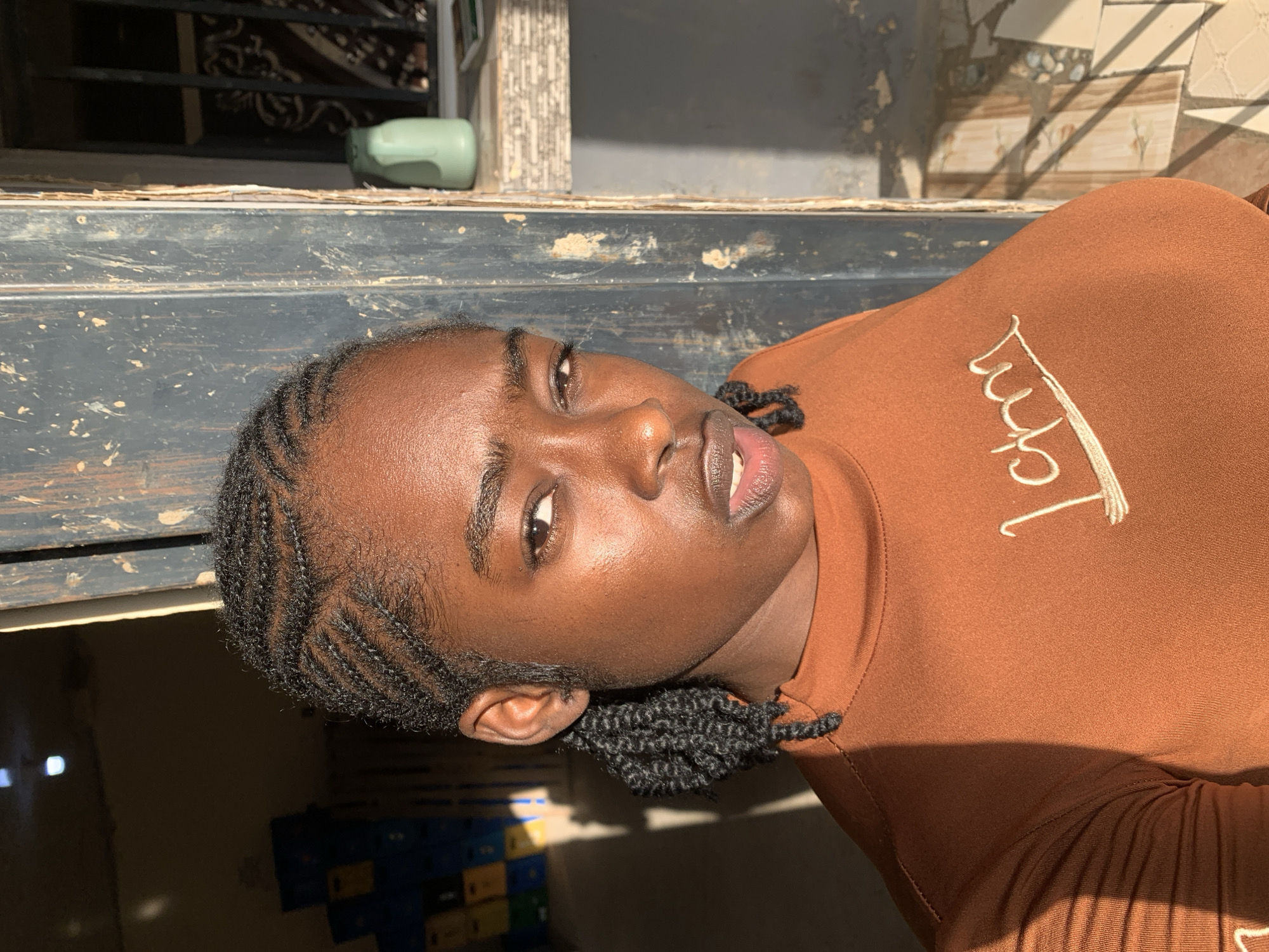 An individual with dark skin and dark hair styled in cornrows that transition into twisted braids on the sides is centrally positioned within a doorway or opening. The person wears an orange-brown long-sleeved top with light-colored embroidered text across the chest. The gaze is directed forward towards the viewer, with a neutral to serious expression and slightly parted lips. Strong, direct sunlight from the right illuminates the right side of the individual's face and upper body, creating distinct highlights and shadows, while the left side is predominantly in shadow.
The background on the left reveals a dark interior space with stacked blue, yellow, and white plastic containers visible in the mid-ground. The background on the right features a dark, weathered vertical door frame or panel. Beyond this, a window with dark security bars is visible, and a light green pitcher along with a small, unidentifiable object are situated on the window sill. The floor on the right side of the scene is composed of light-colored, geometrically patterned tiles. This scene is located in the City of New Stadium, Nigeria.