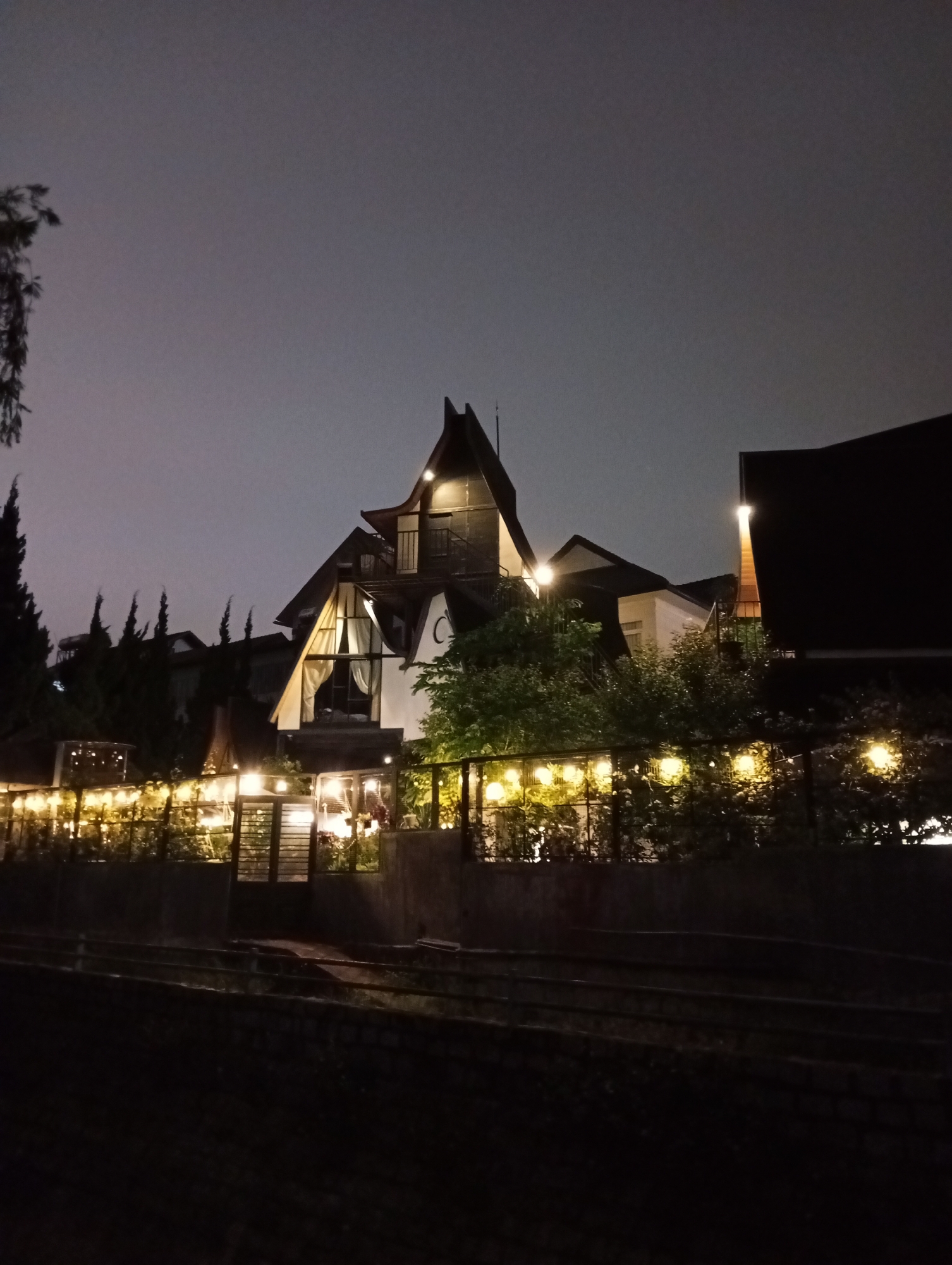 The image presents a multi-story building at night in Xuân Hương, Đà Lạt, Vietnam. The night sky is a dark, uniform grey. The building exhibits a distinctive architectural style characterized by multiple sharply angled gables and steeply pitched rooflines of a dark color. Artificial lights with a warm color temperature illuminate the structure, highlighting balconies, upper levels, and a lower-level window fitted with white curtains. In the mid-ground, a dark fence composed of horizontal slats, featuring a central gate, is adorned with multiple strings of spherical, warm-toned lights. Various evergreen trees and dense bushes are situated around the building and behind the fence, with some foliage catching the illumination. A tall, slender tree is partially visible on the far left. The foreground consists of a dark, uneven ground surface, possibly a path or terraced section, bordered by a low, dark retaining wall. There are no individuals visible within the scene.