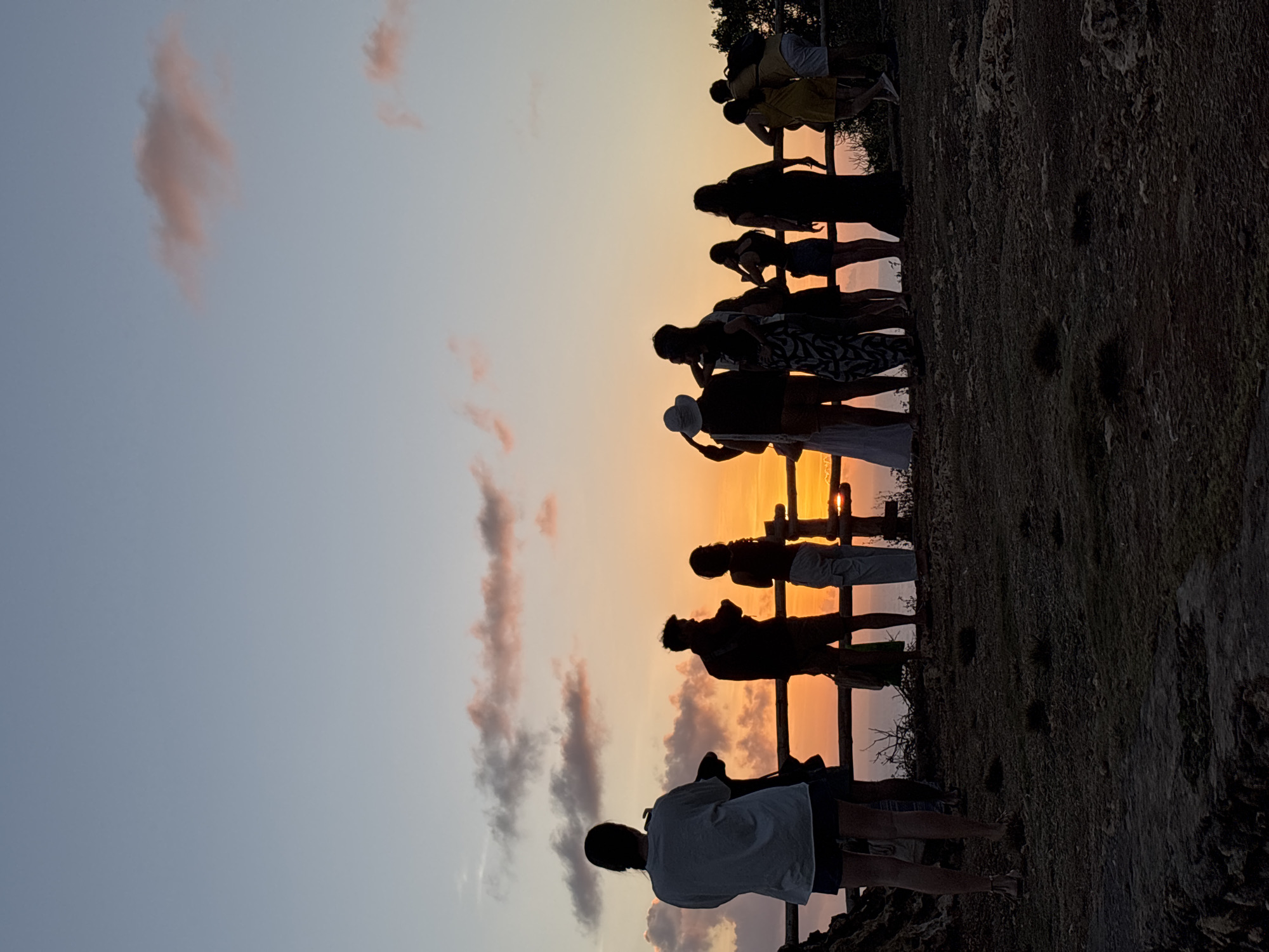 A group of people are silhouetted against a sunset on a cliff overlooking the ocean. The sky is a gradient of pale blue and orange, with soft pink clouds. A wooden fence runs horizontally across the frame, and the people are gathered behind it, looking out at the view. The ground in the foreground is rocky and uneven, with sparse vegetation. The group appears to be a mix of ages and genders, some standing close together while others are more spread out. The overall atmosphere is peaceful and serene, with a sense of awe as the sun sets over the horizon.