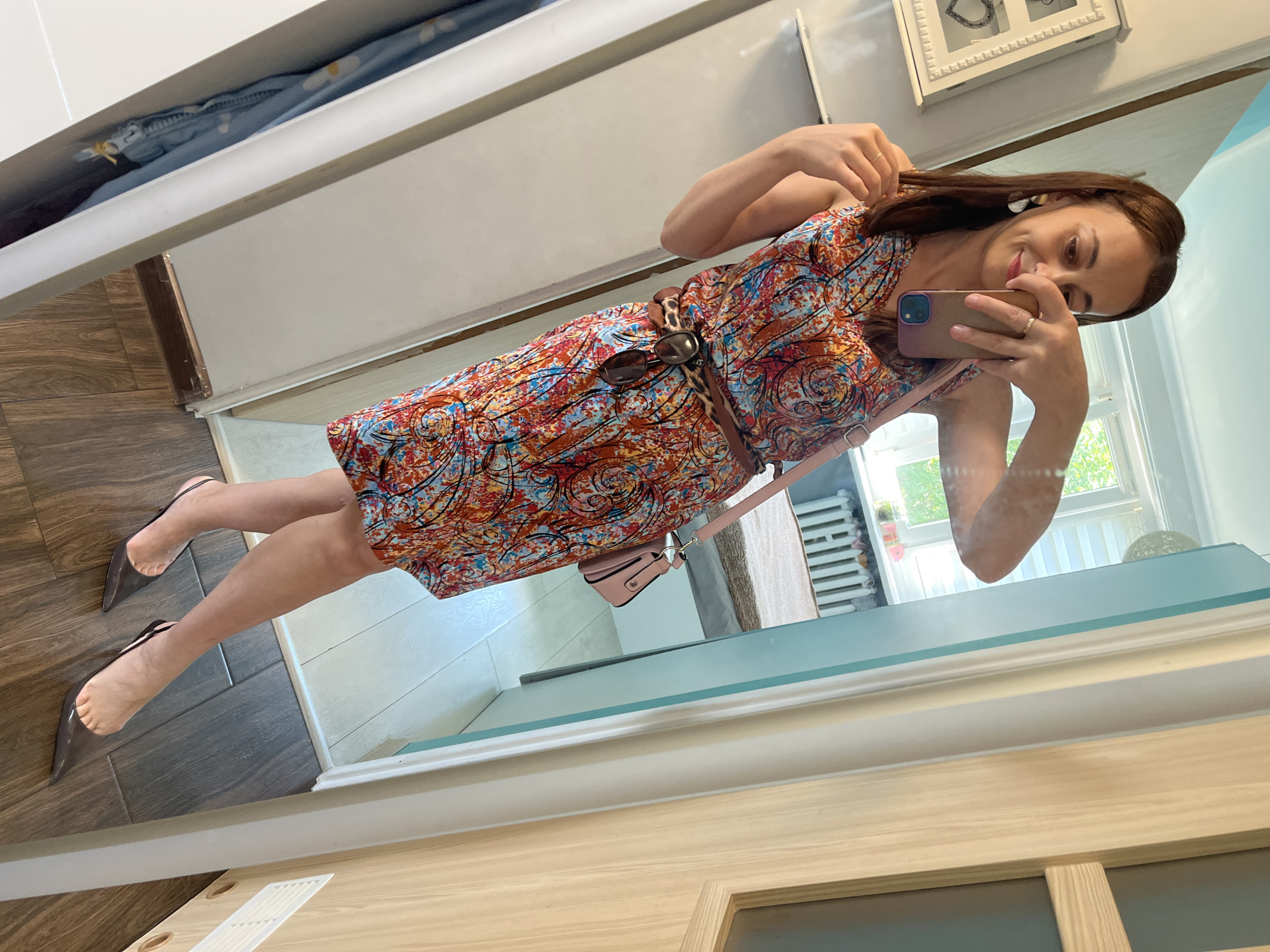 A woman stands in a room, taking a selfie in a mirror. She is wearing a sleeveless, v-neck, knee-length dress with a colorful, abstract print. The dress is cinched at the waist with a brown belt that has leopard print accents and hangs a pair of sunglasses. She is also carrying a small pink handbag. The woman has on clear pointed-toe slingback heels. She is holding her phone in her right hand, and her left hand is touching her hair. The background of the mirror shows a window with blinds, a radiator, and a bed. The room has wooden flooring and a white doorframe.