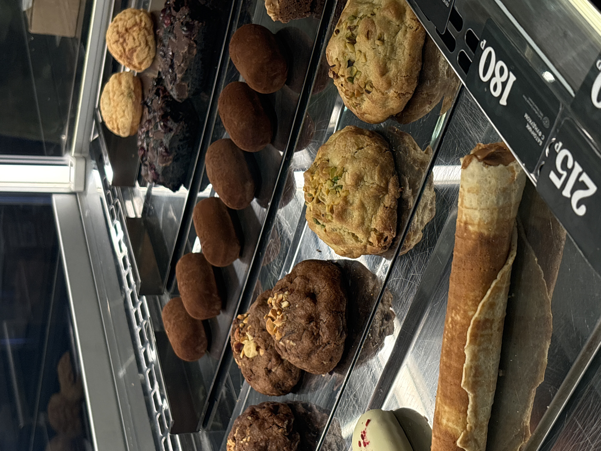 A display case in Borovoye Matyushino, Russia contains several types of desserts: round cookies, chocolate pastries, oval-shaped brown candies, pistachio cookies, chocolate cookies with nuts, and a rolled crepe. Price tags of 180 and 215 are visible on the right.