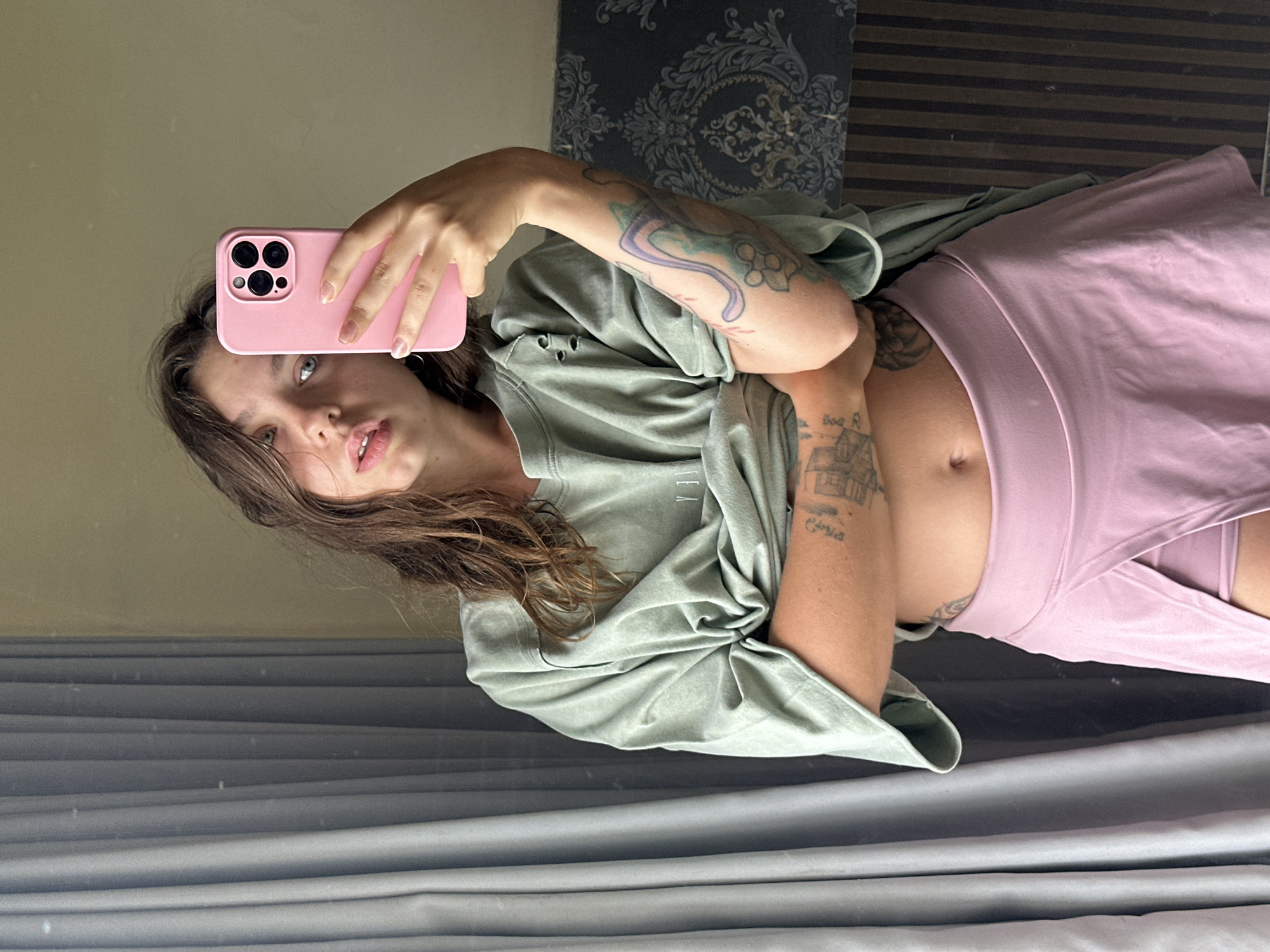 A young woman with brown hair takes a mirror selfie indoors. She is wearing an oversized, light green t-shirt that she has pulled up to expose her midriff, and a pink skirt. She has tattoos on her arms and torso, and is holding a pink smartphone in her right hand. The background includes a grey curtain on the left, a wall with a patterned wallpaper and vertical stripes behind her, and a beige wall to the right. The lighting appears to be natural, coming from a window.