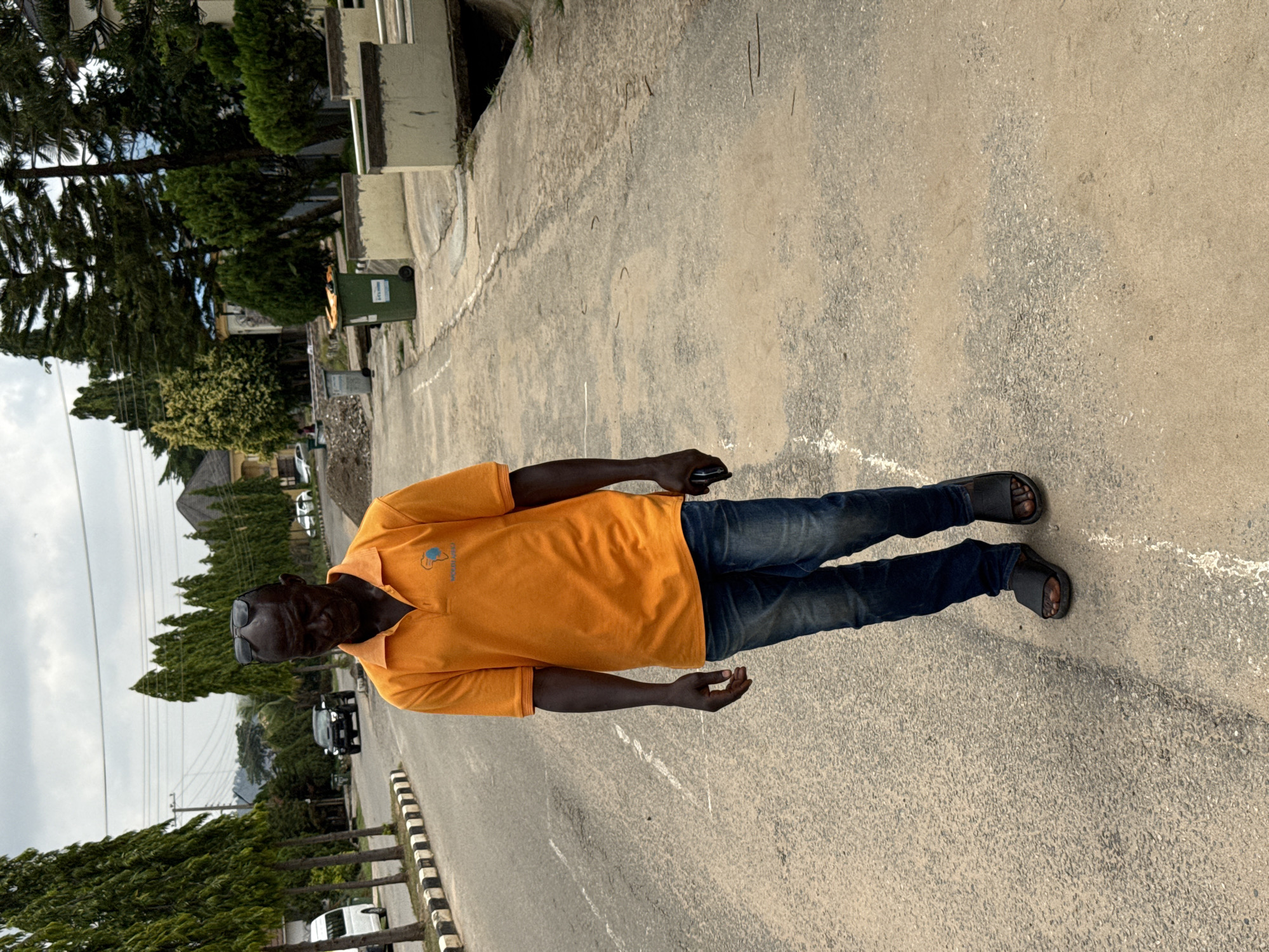 A man stands on a gray asphalt road in Abuja, Nigeria. He wears a bright orange polo shirt, dark denim jeans, and dark-colored sandals. He has dark skin and wears dark sunglasses. In his right hand, he holds a dark object. The road is bordered by a black and white striped curb and rows of trees. Behind him, there are houses and vehicles. A green waste bin is visible near a building. The sky is overcast.
