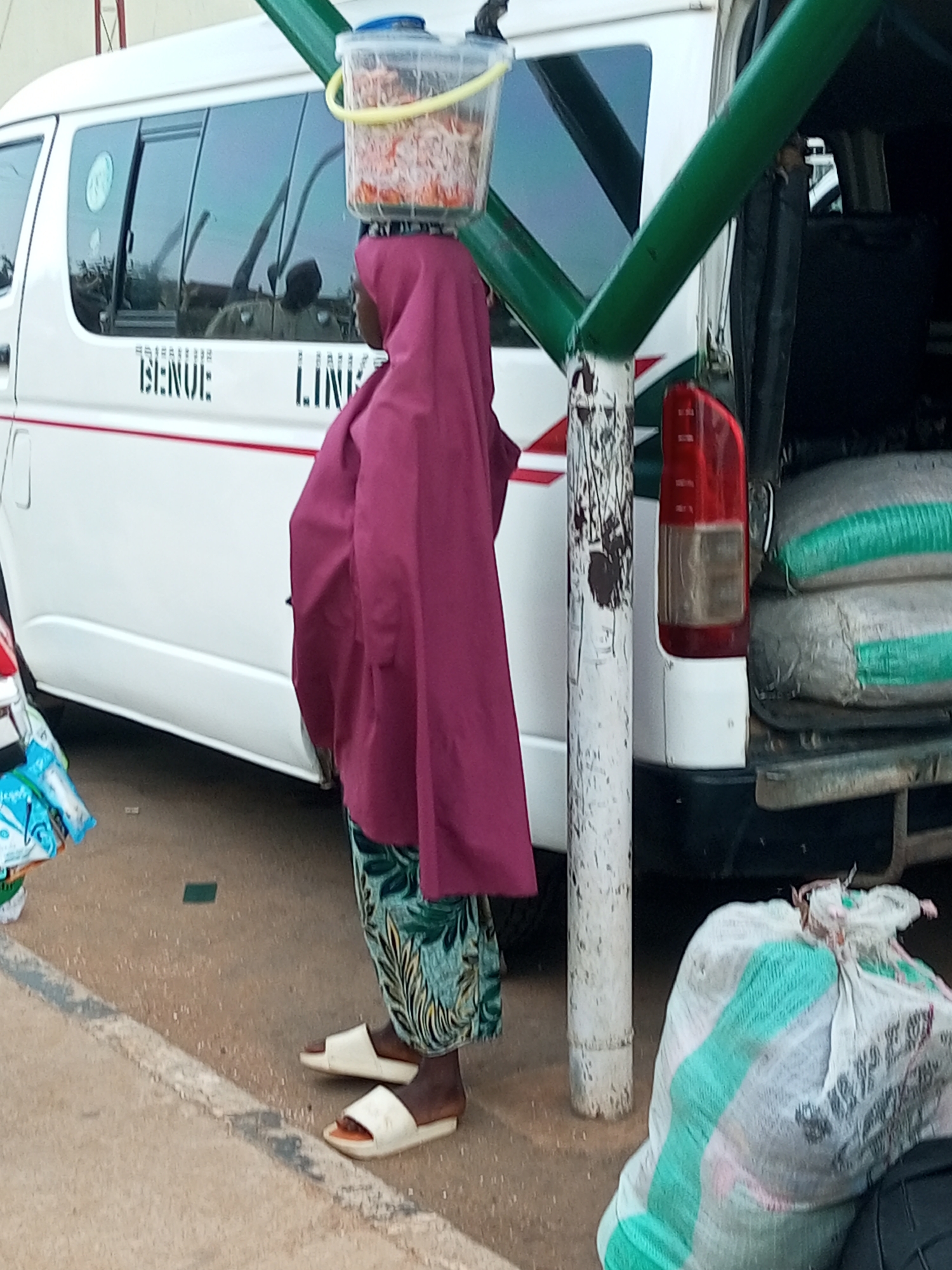 A woman balances a clear plastic container of food on her head while standing next to a white minibus with "BENUE LINE" written on its side. She is wearing a purple headscarf and a patterned skirt, and white sandals. The minibus appears to be parked, with its rear doors open, revealing bags of goods inside. In the foreground, a large white sack with green stripes is visible. The scene takes place outdoors on a dirt or gravel surface, likely in an urban setting. The overall impression is of a street vendor or someone engaged in commerce.
