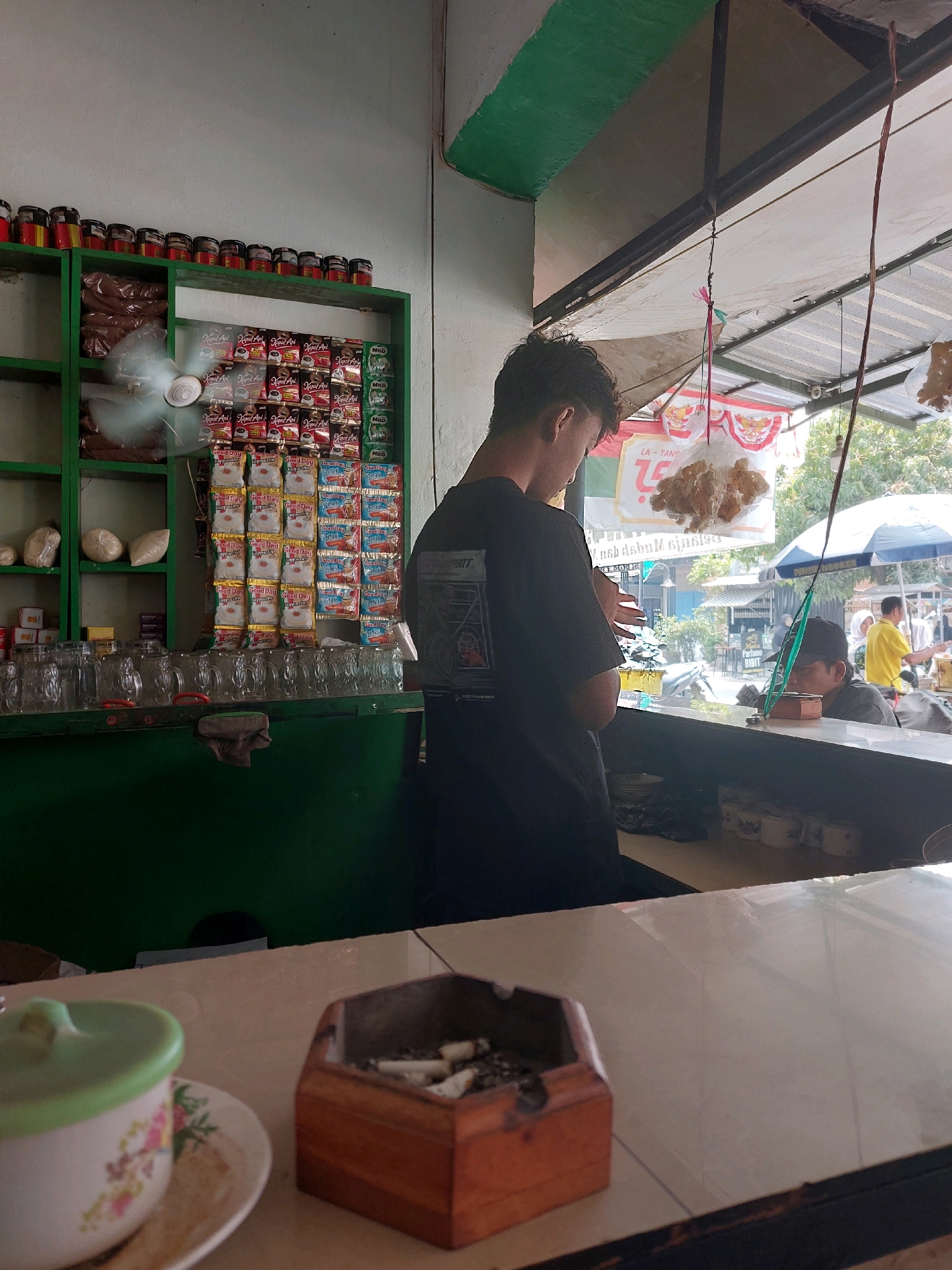 The image depicts a street-side food stall in Sumuragung, Indonesia. In the foreground, a wooden ashtray filled with cigarette butts sits on a light-colored counter. To the left, a white ceramic cup with a green lid and floral design rests on a saucer. Behind the counter, a young man in a black t-shirt is seen from the back, seemingly preparing or serving something. Shelves behind him are stocked with various products, including bags of coffee and other packaged goods. A small fan is visible on one of the shelves. In the background, other people are present, some seated, and a few are visible through an open awning, suggesting an outdoor seating area. The overall scene conveys a casual and functional atmosphere typical of a local Indonesian eatery.