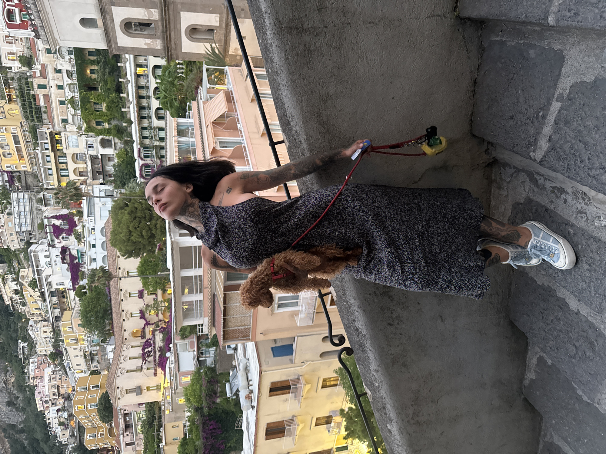 In Arienzo, Italy, a woman with extensive tattoos stands on a stone staircase, holding a small brown poodle. The woman is wearing a dark, polka-dotted sleeveless dress and light blue sneakers. Her eyes are closed, and her head is tilted back. The poodle is looking to the right. A red leash, attached to the dog, dangles from the woman's hand, with a small yellow object hanging from it. Behind the woman, a hillside is covered with densely packed buildings in various shades of yellow, white, and pink, characteristic of Amalfi Coast architecture. Lush green foliage and purple flowers are visible among the structures. A tall bell tower is partially visible on the right side of the frame. The lighting suggests it is late afternoon or early evening.