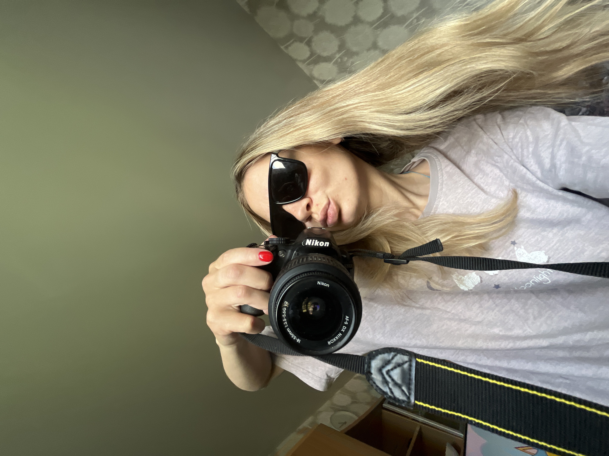 A young woman with long blonde hair, wearing sunglasses and a pink t-shirt, takes a selfie with a Nikon camera. She is puckering her lips and holding the camera up to take the photo. The background appears to be an indoor setting with patterned wallpaper. The image does not provide any information about the location in the city of Chaikovsky, Russia.
