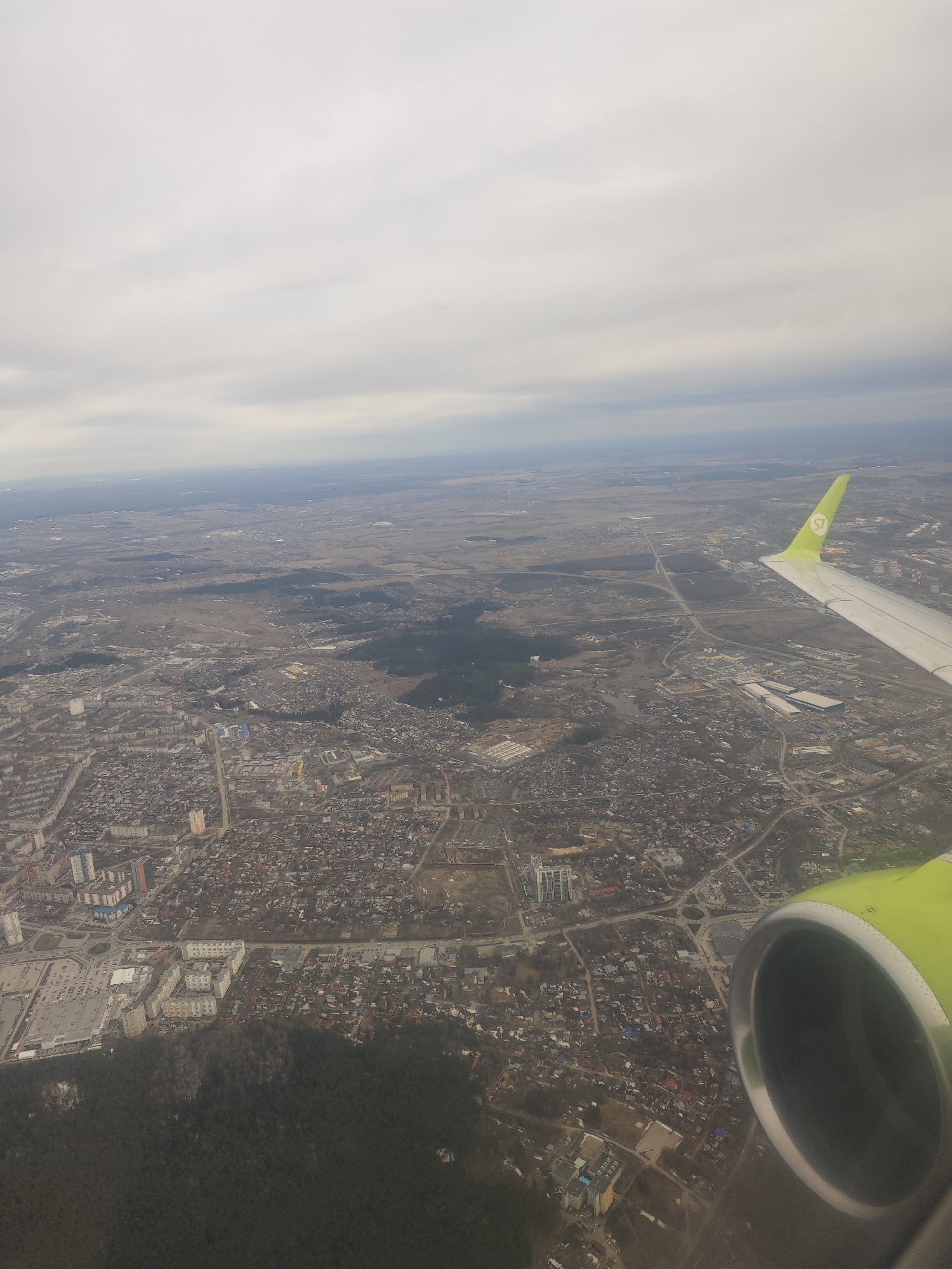The image shows an aerial view from a plane, presumably during descent or ascent, over the city of Novosibirsk, Russia. An aircraft wing and engine are visible in the foreground, with the "S7" logo on the wingtip. Below is a densely populated urban area with a mix of residential and industrial buildings. Forested areas are interspersed within the city. In the distance, a more rural landscape is visible under an overcast sky. The image captures a typical cityscape transitioning into surrounding countryside.