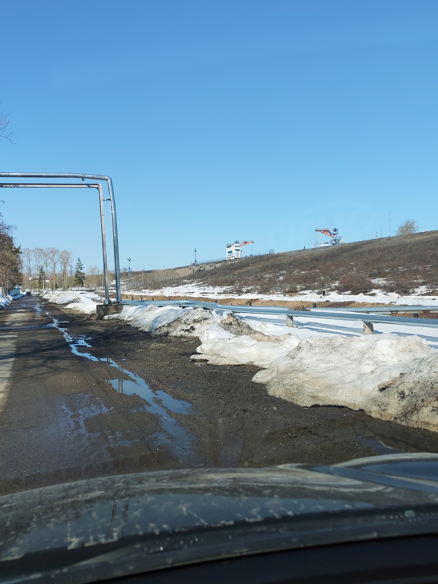 View from inside a vehicle driving on a wet, muddy road with remnants of melting snow on either side. The road appears to be adjacent to a raised embankment or slope, with metal guardrails running along the edge. On top of the embankment, a structure is visible, and behind it, two cranes with orange booms stand against a clear blue sky. A metal frame or archway structure stands on the left side of the road. The city is Chaikovsky, Russia.