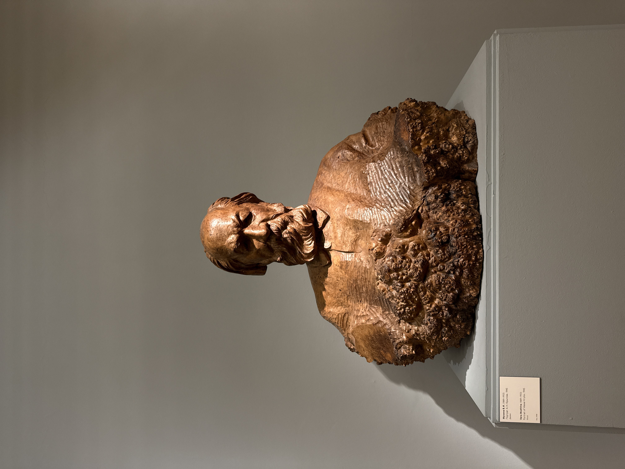 The image shows a wooden bust sculpture of a man with a beard, likely A.N. Krylov, created by Vera Mukhina in 1945. The sculpture is displayed on a wall, potentially in a museum or gallery. The bust is a textured brown color. The scene appears to be indoors, and the background wall is a neutral gray color. There is a small informational plaque next to the sculpture. The text on the plaque indicates that the bust is a portrait of A. N. Krylov, created by Vera Mukhina in 1945, and that the medium is wood. The location in Moscow, Russia, is implied as being a museum or gallery.