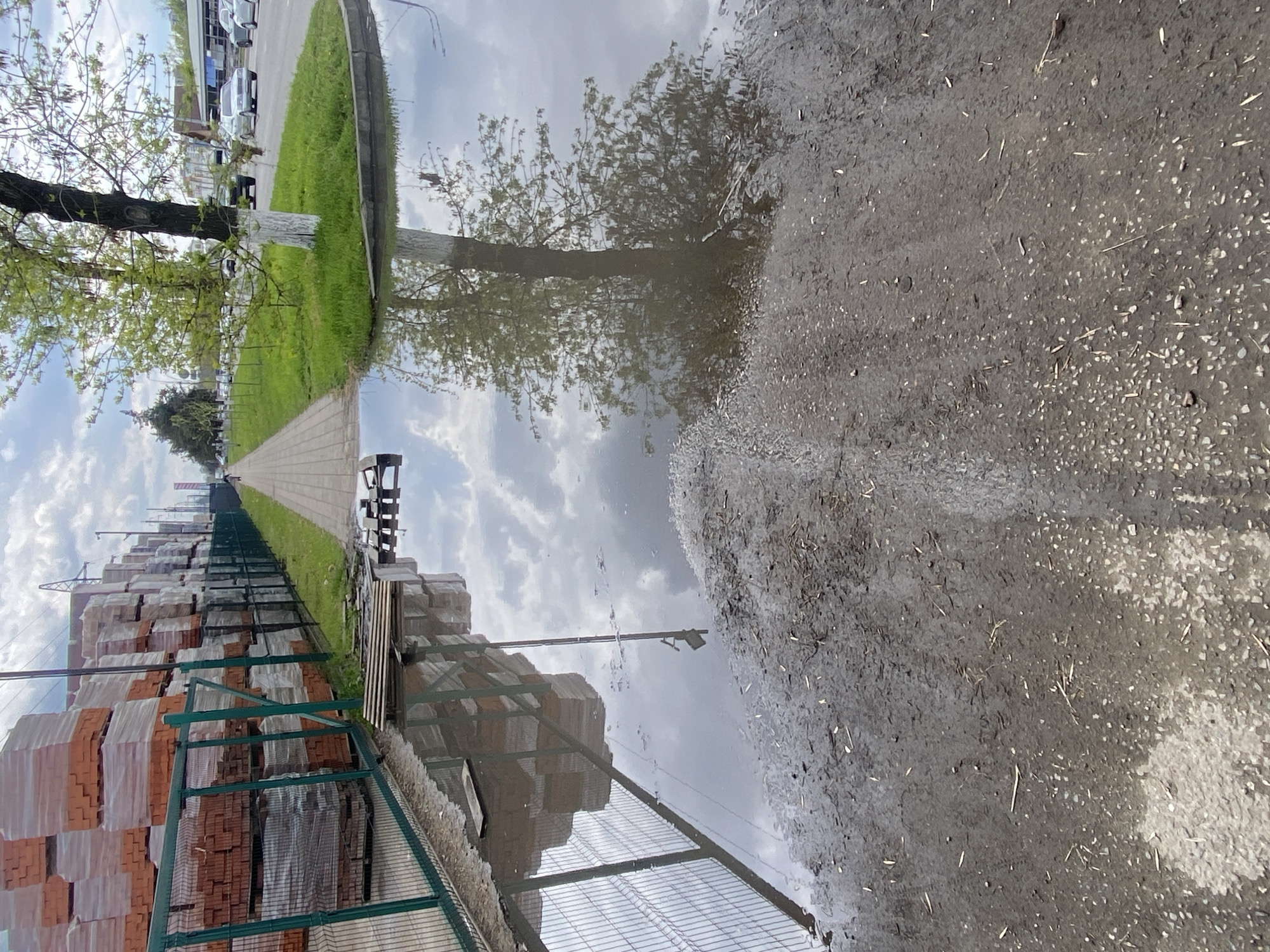 The image depicts a puddle reflecting a scene of a construction supply yard, likely following rainfall. Stacks of wrapped red bricks are visible behind a green wire fence, bordering a sidewalk with green grass and trees beyond. On the other side of the puddle, a muddy ground is visible. The reflection shows the sky with clouds and a tree. The location is in the city of Unknown, Unknown.
