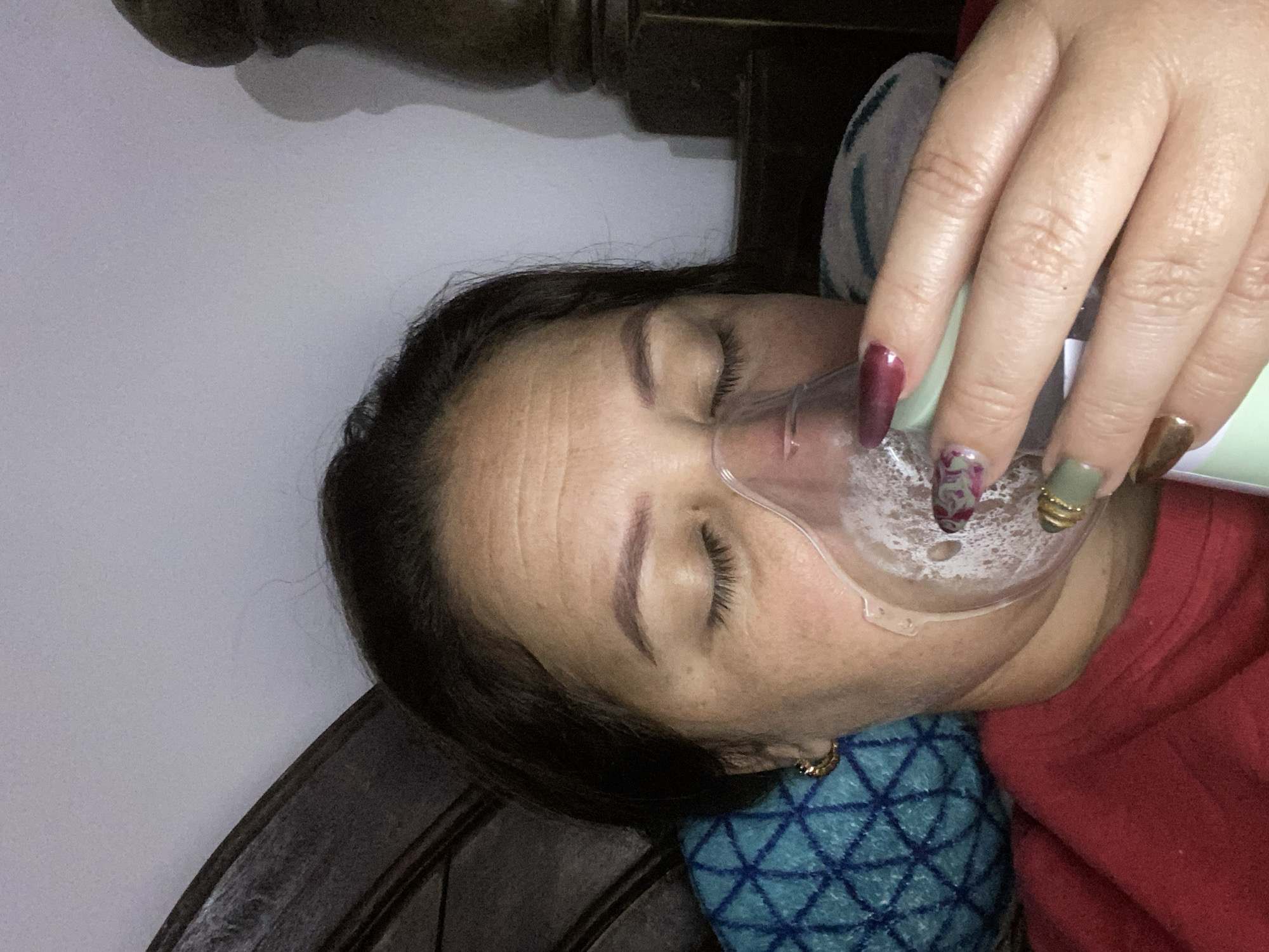 A close-up shot shows a woman with her eyes closed, wearing an oxygen mask. Her head is resting on a blue, patterned cushion. The mask covers her nose and mouth, and appears to be connected to an oxygen source, though the source itself is not visible. The woman has dark hair and her skin appears to be light-toned. She is wearing a red shirt. Her right hand is visible in the upper right corner of the frame, with long, painted fingernails in shades of burgundy, green, and gold. The background is softly lit, with hints of dark wood and a light-colored wall. The overall impression is of someone resting or receiving medical care. There is no visible text in the image.